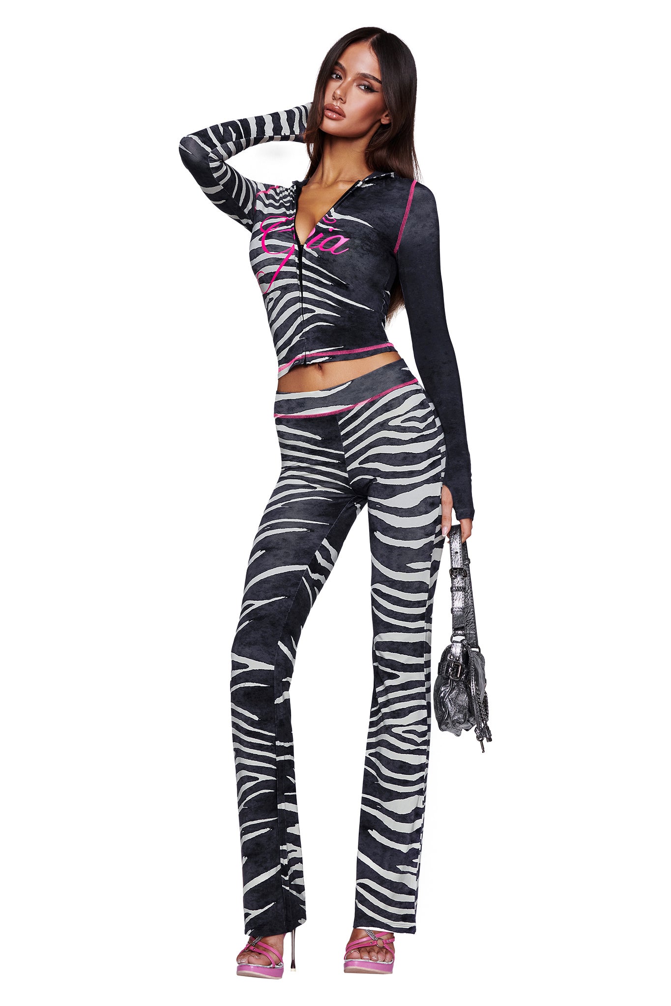 A woman poses in the I.AM.GIA BLARE TRACKPANT - BLACK WILD SIDE, styled with a coordinating long-sleeve top and matching bag. She completes the bold zebra print look with pink heels, standing confidently against a white background.