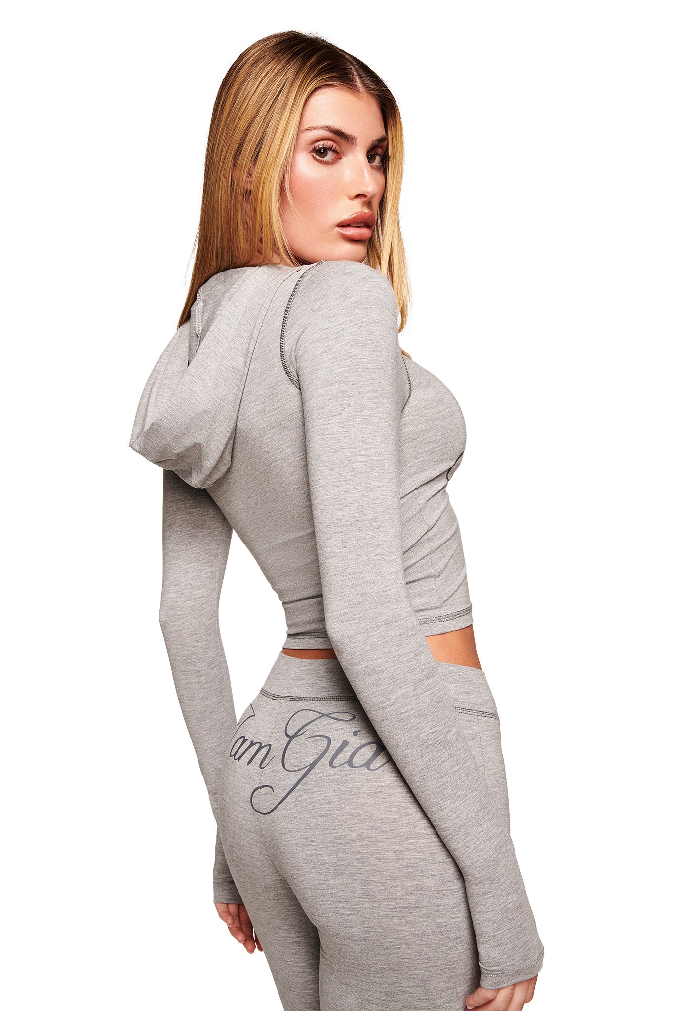 A woman with long blonde hair wears the fitted BLARE JERSEY HOODIE in heather grey by I.AM.GIA, paired with matching pants featuring the brand’s name on the back, as she stands sideways looking over her shoulder.