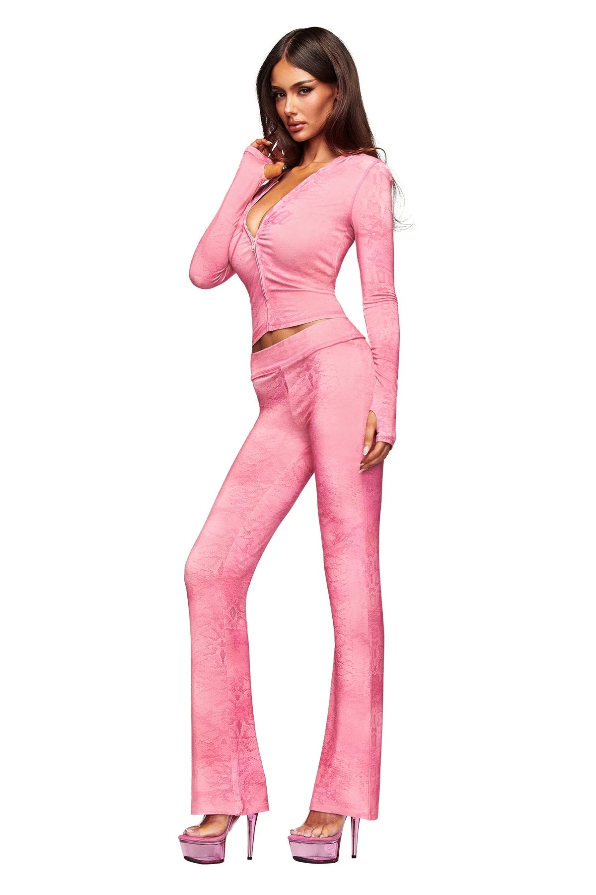 A woman stands against a white background wearing the I.AM.GIA BLARE TRACKPANT in Light Pink Serpentine with a flared leg, styled with a fitted pink velour zip-up top and pink platform heels.