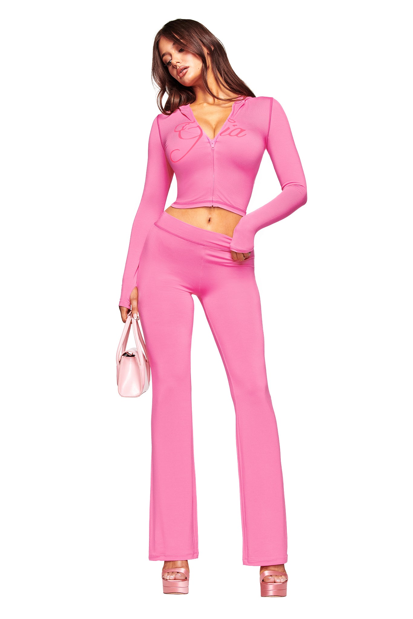 A woman in the I.AM.GIA BLARE HOODIE - NEON PINK poses against a white background, styled with matching flared pants, a shiny pink handbag, and pink platform heels.