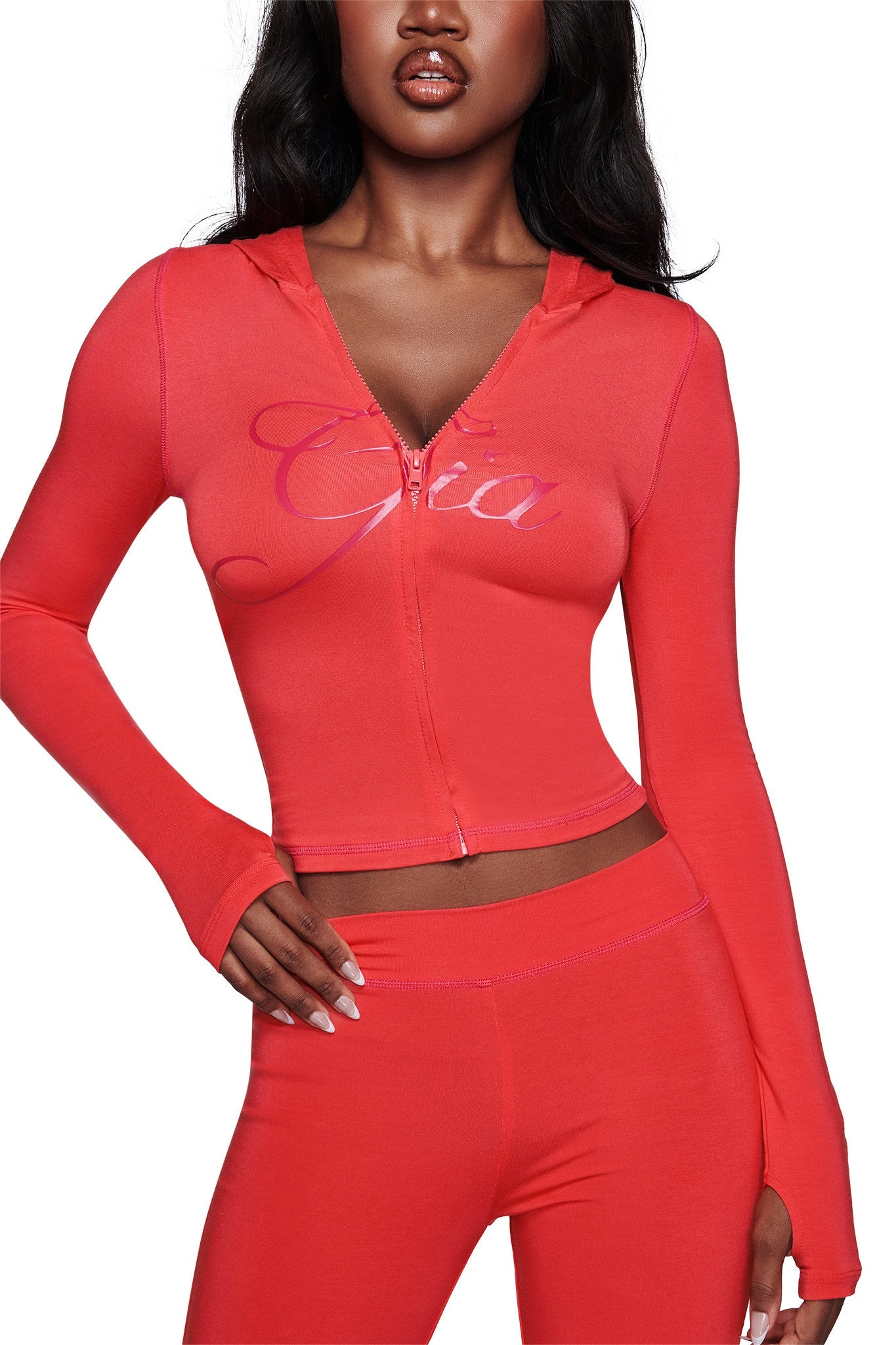 A woman wears the fitted, bright papaya BLARE HOODIE by I.AM.GIA, a zip-up style with long sleeves and thumb holes. "Gia" is scripted across the chest as she poses confidently with one hand on her hip.