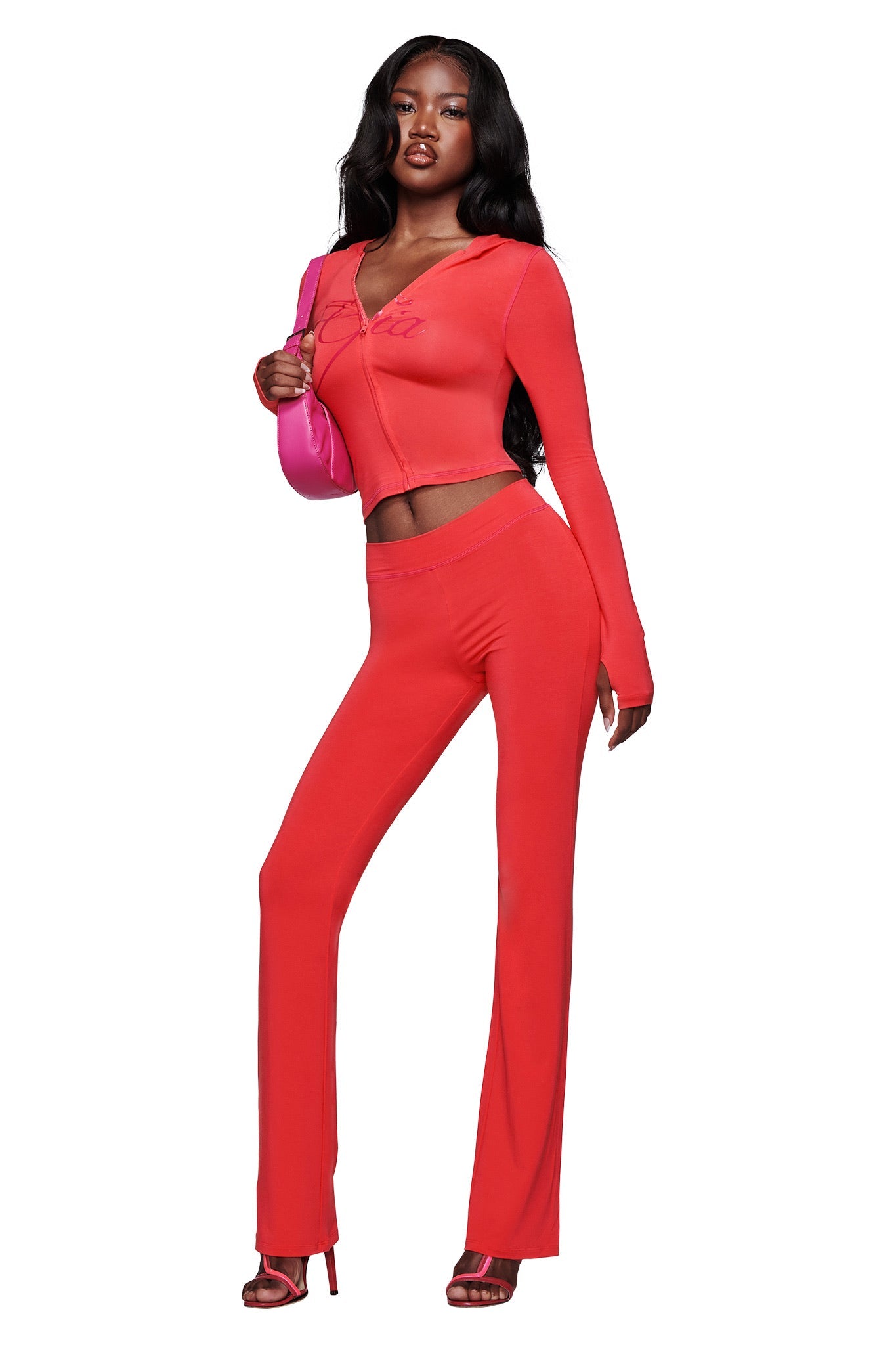 A woman stands confidently in the I.AM.GIA BLARE TRACKPANT - PAPAYA, a fitted, bright red two-piece with a mid-rise waist and flared leg. She accessorizes with a pink shoulder bag and matching red heels against a white background.