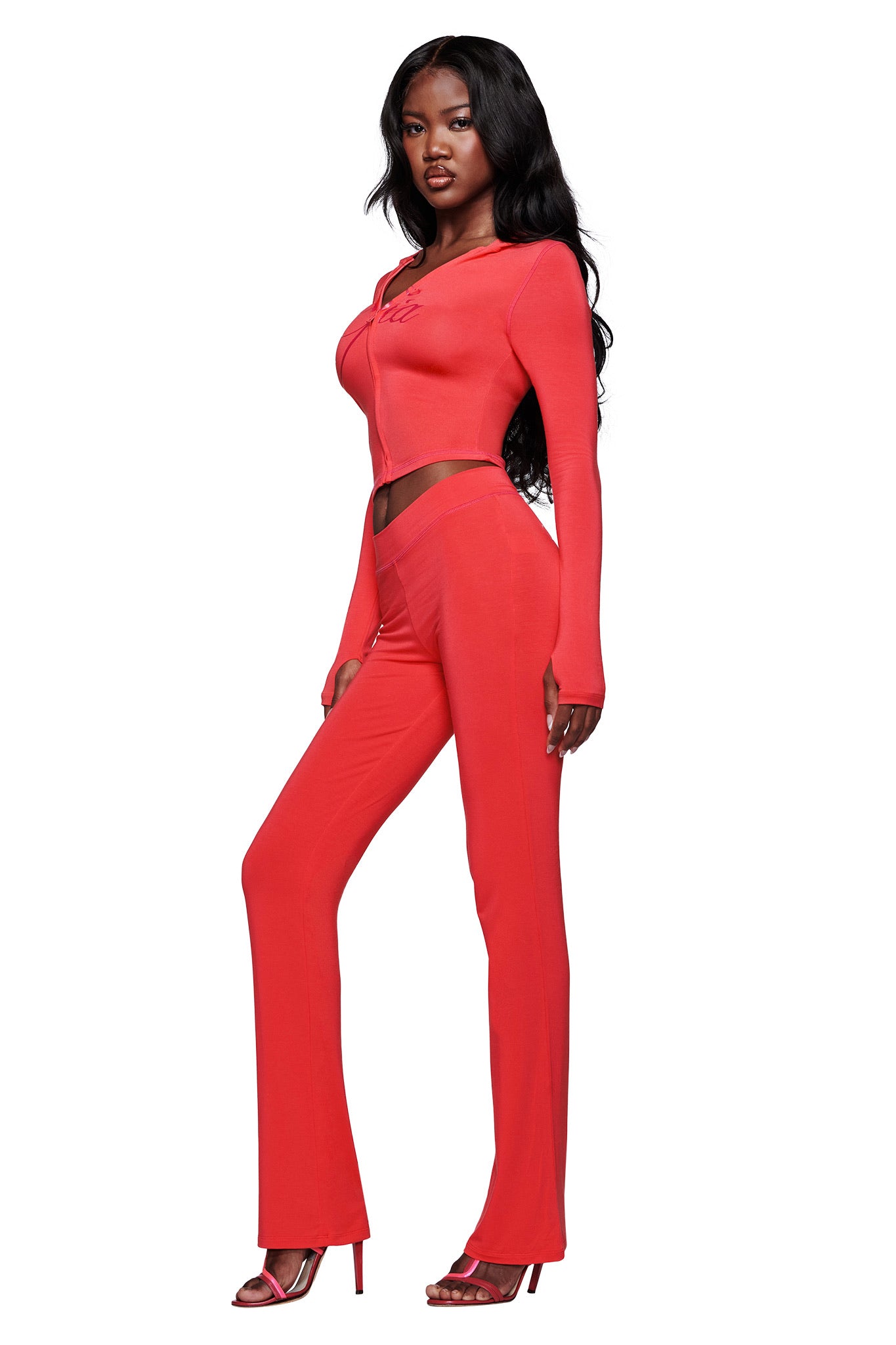A woman with long black hair poses in a bright red two-piece set with long sleeves and flared pants, layered under an open I.AM.GIA BLARE HOODIE in Papaya, paired with matching high-heeled sandals against a plain white background.
