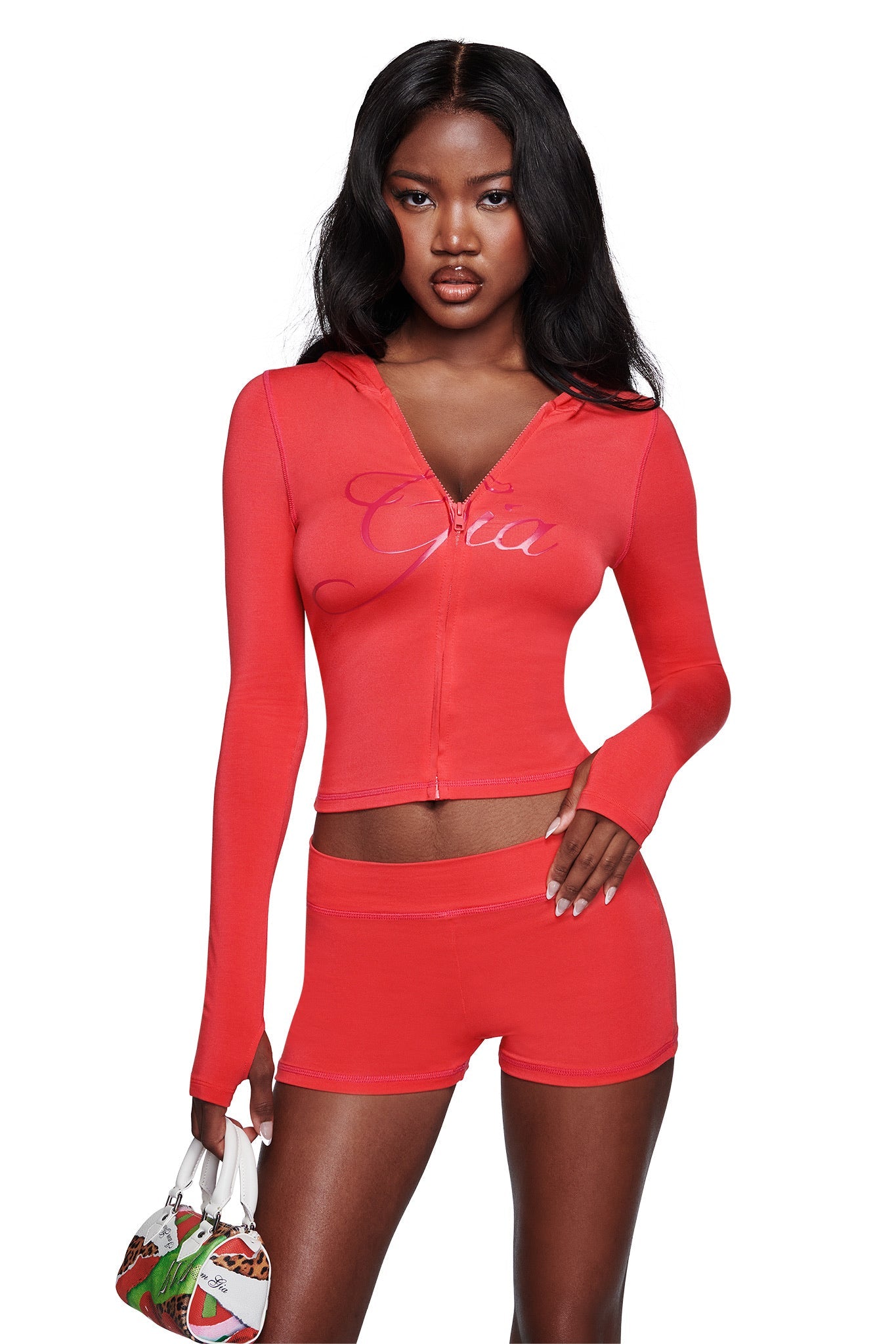 A woman in a fitted, bright red zip-up crop hoodie and I.AM.GIA BLARE SHORT - PAPAYA with an elastic waistband holds a colorful handbag. She has long, wavy dark hair and stands against a plain white background.
