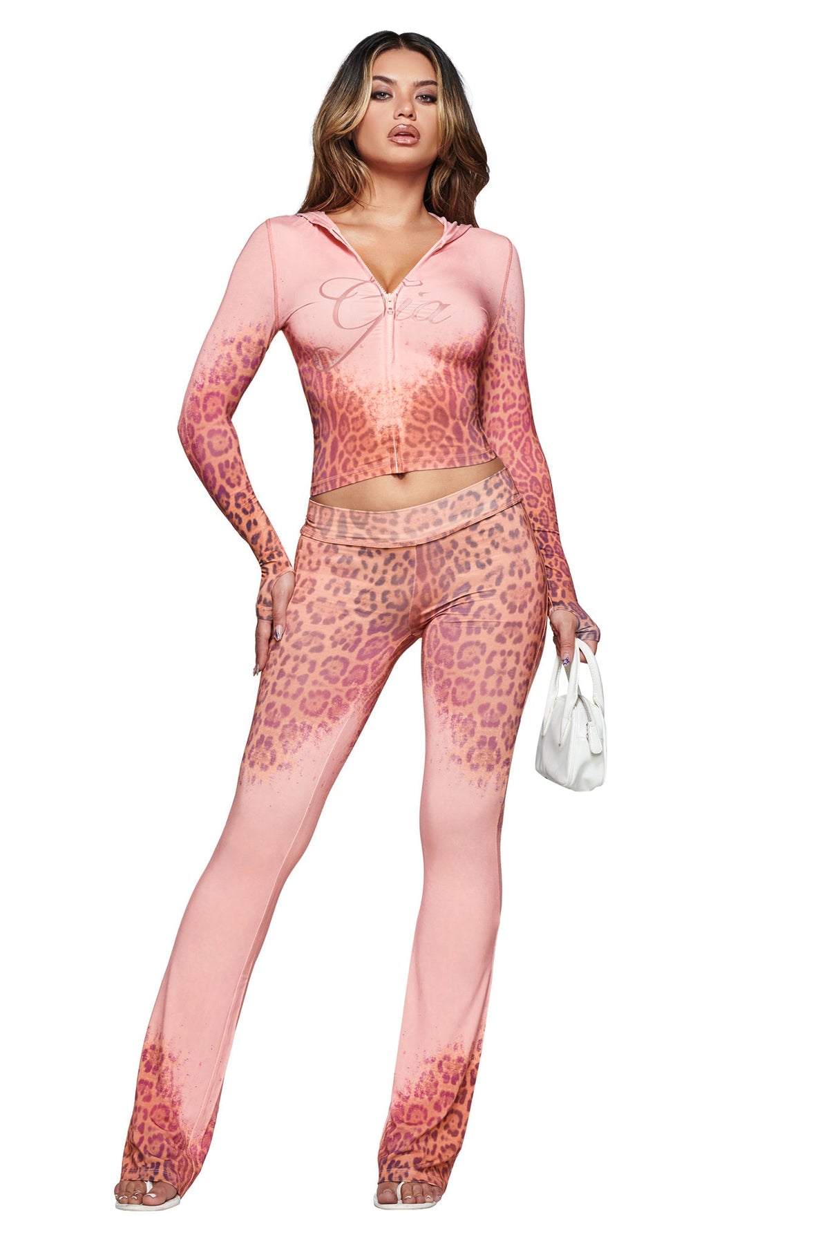 A woman wearing the I.AM.GIA BLARE HOODIE in pink faded panthera leopard print and matching flared pants, holding a small white handbag, stands against a white background.
