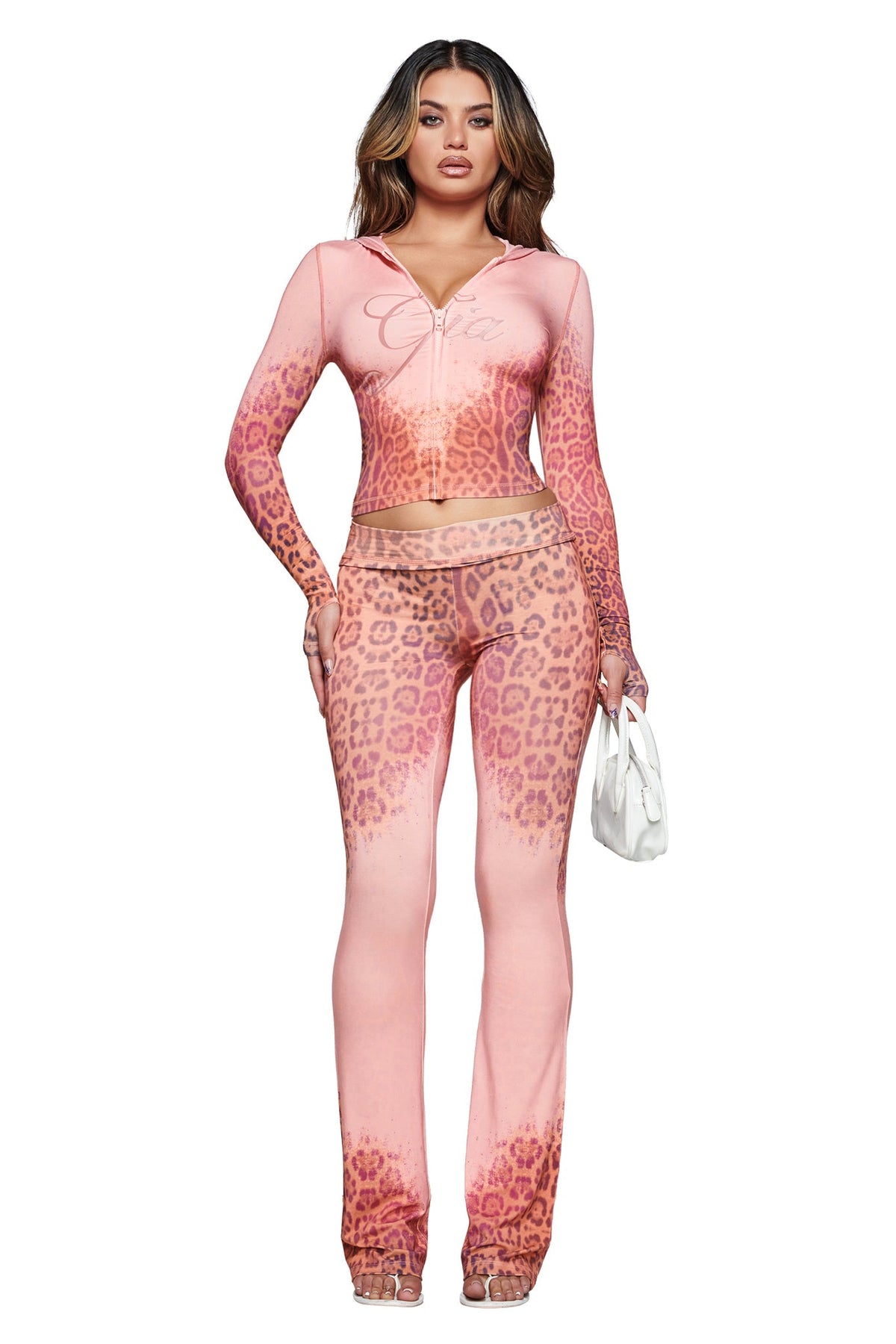 A woman stands facing forward, wearing the BLARE HOODIE in Pink Faded Panthera from I.AM.GIA paired with clear heels, and holding a small white handbag.