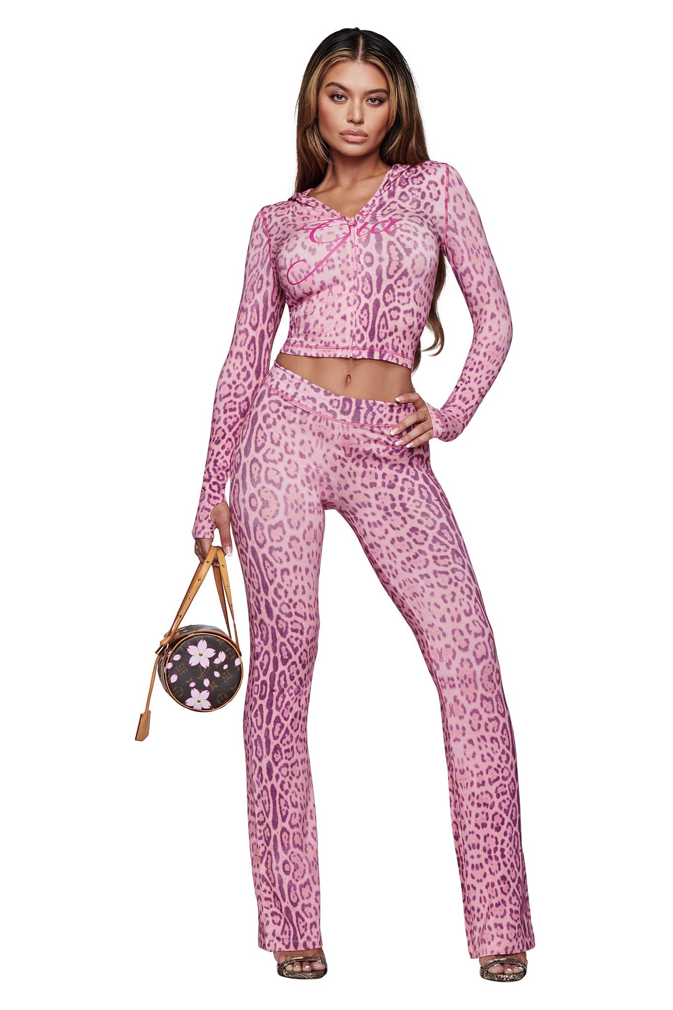 A woman stands confidently in I.AM.GIA’s BLARE TRACKPANT - PINK PANTHERA, paired with a matching top and high heels, holding a round floral handbag against a white background.