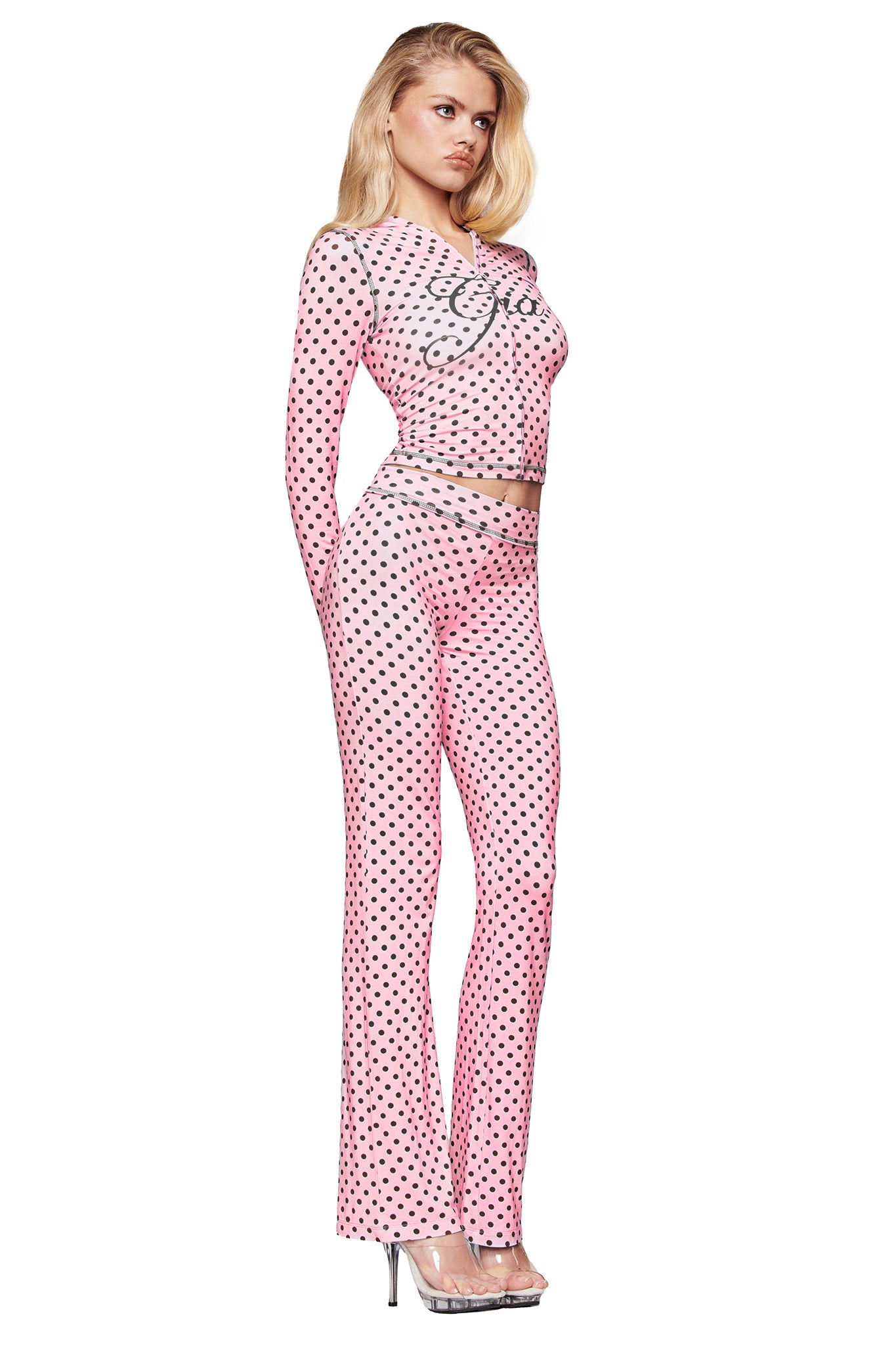 A woman with long blonde hair models the I.AM.GIA BLARE HOODIE - PINK POLKA DOT, a fitted, long-sleeve light pink hoodie with black polka dots, styled in profile and paired with clear high-heeled platform shoes.