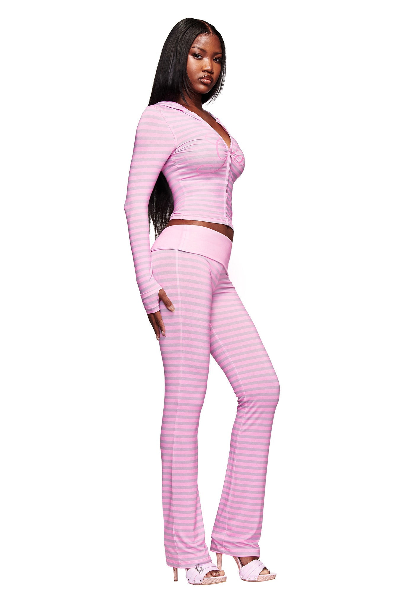 A woman poses confidently in the I.AM.GIA BLARE HOODIE - LIGHT PINK STRIPE with matching high-waisted pants and open-toed heels, her long straight hair framing her face against a white background.