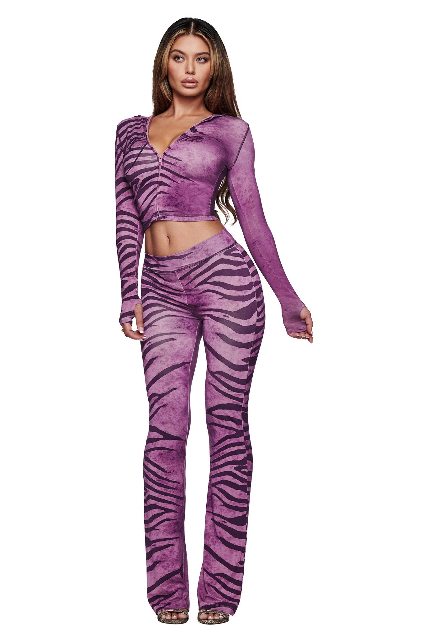A woman with long hair and heels faces forward against a white background, wearing the I.AM.GIA BLARE TRACKPANT - PLUM WILD SIDE, featuring a bold purple and black zebra stripe print with a flared leg and matching long-sleeve cropped top.