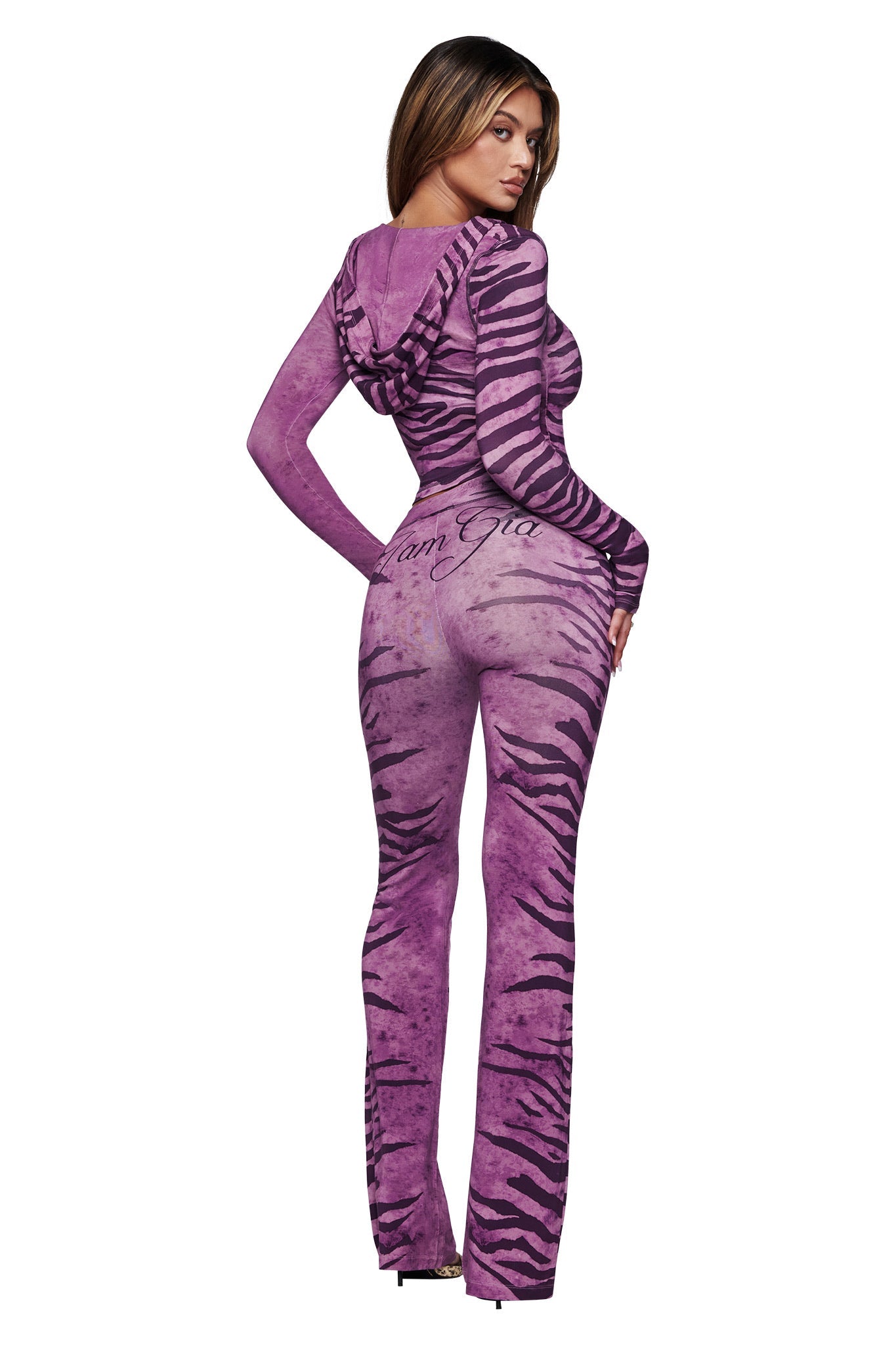 A woman with straight long hair poses against a white background, facing away and looking over her shoulder. She wears I.AM.GIA's BLARE TRACKPANT in the Plum Wild Side print, featuring a fitted silhouette and flared legs.