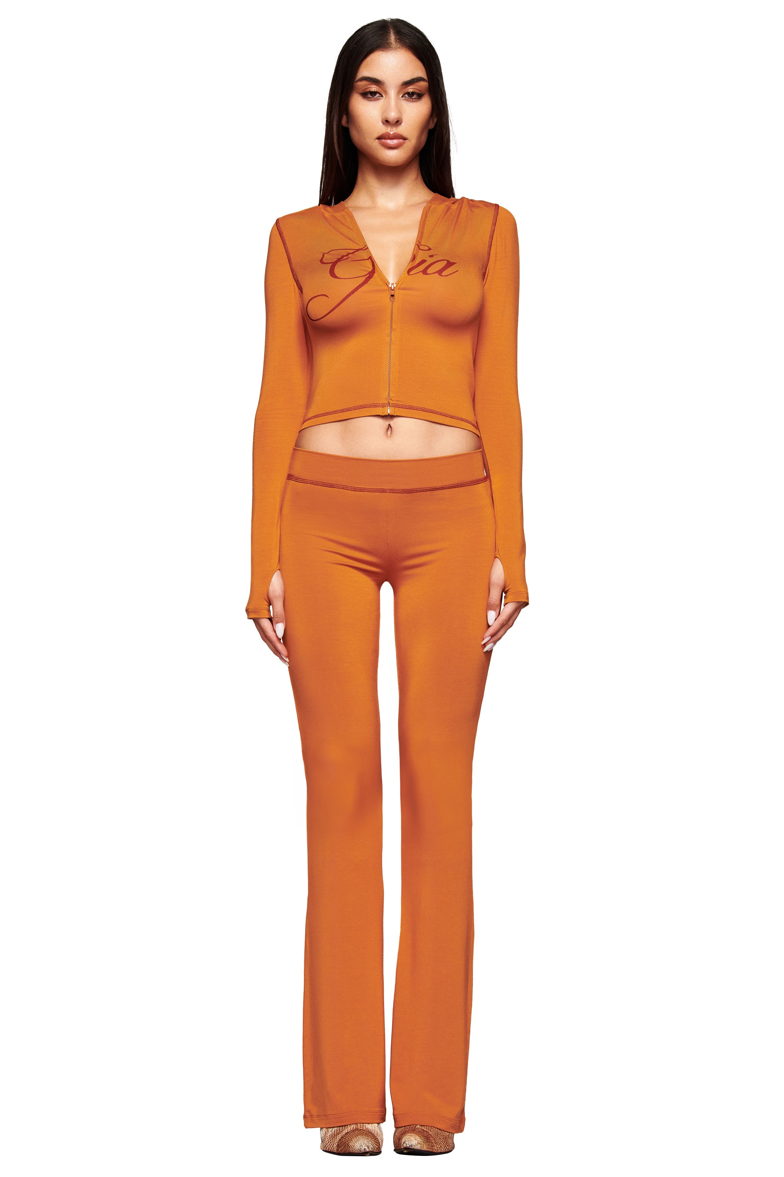 A woman with long hair stands facing forward against a white background, wearing the I.AM.GIA BLARE TRACKPANT in pumpkin and a matching fitted zip-up crop top. She has a neutral expression.