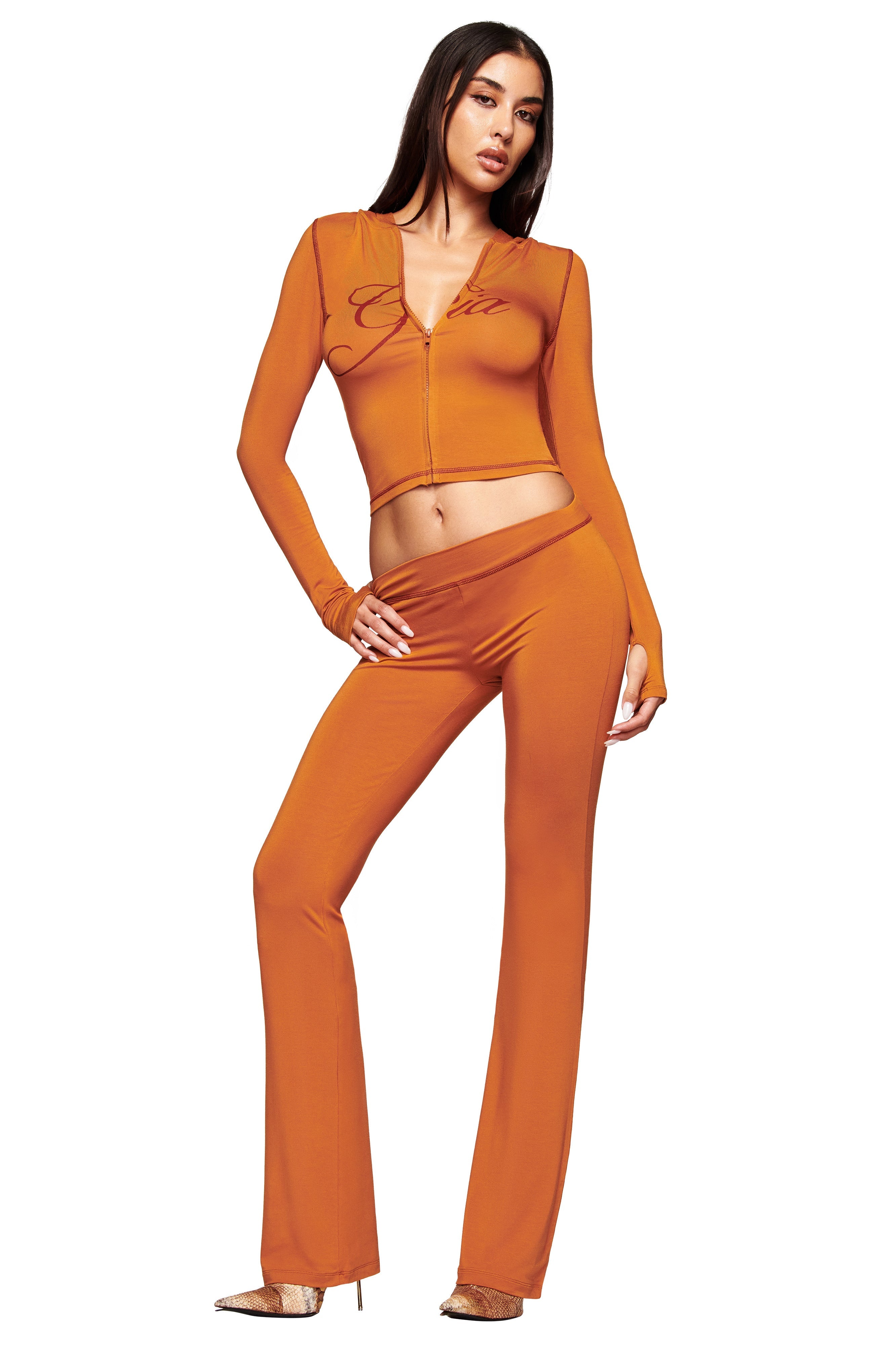 A woman poses confidently against a white background, wearing I.AM.GIA’s pumpkin-colored BLARE TRACKPANT with a low-rise waist and matching zip-up long-sleeve crop top.