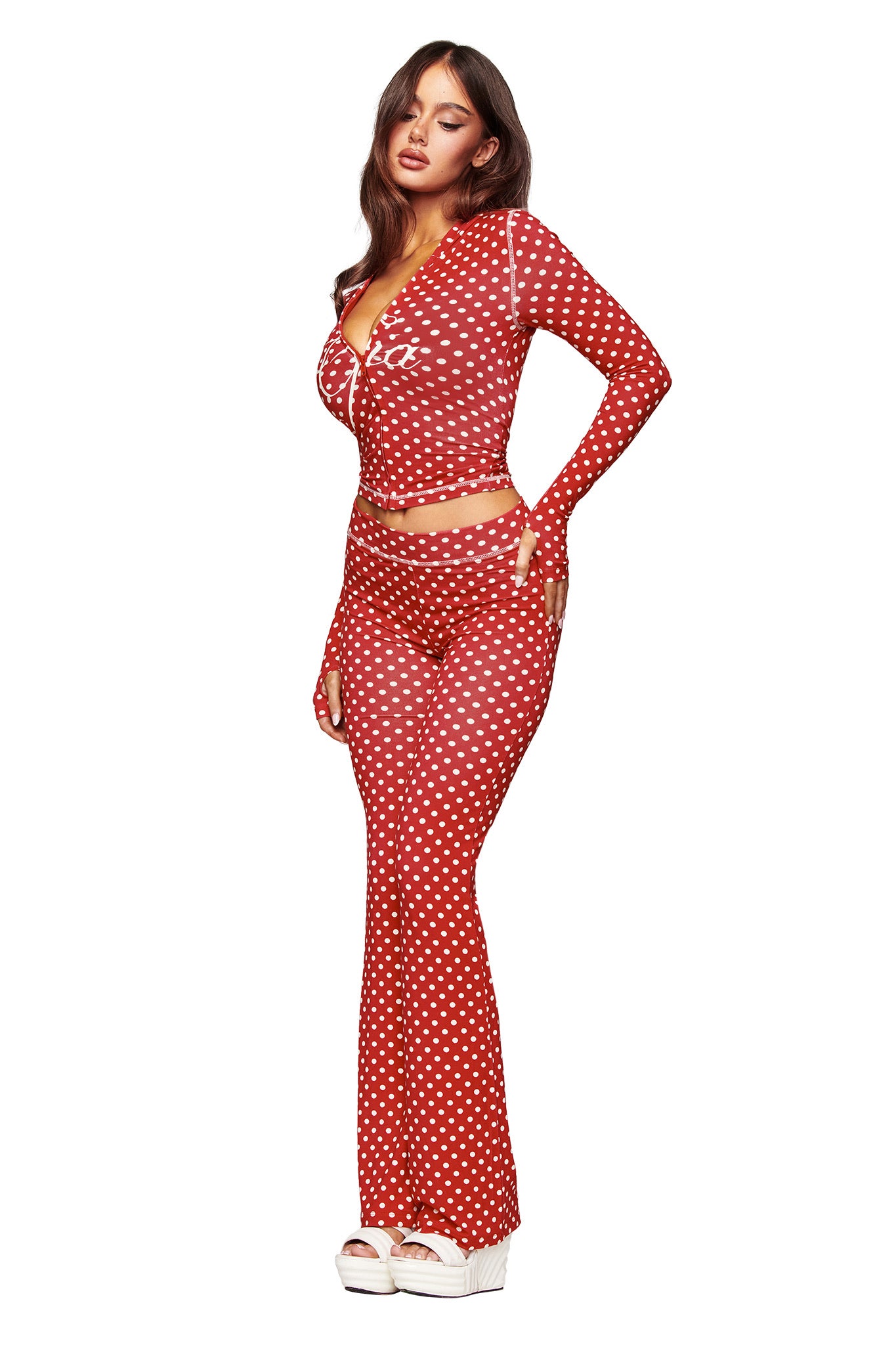 A woman poses against a white background wearing I.AM.GIA’s BLARE TRACKPANT - RED POLKA DOT, styled with a matching long-sleeve fitted top and white platform sandals.