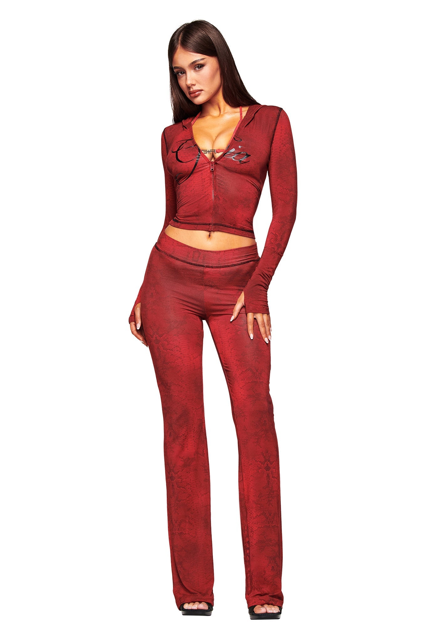 A woman poses against a white background, confidently wearing the I.AM.GIA BLARE TRACKPANT - RED SERPENTINE with a red serpentine print, paired with a fitted red zip-front crop top; both pieces feature subtle patterns.