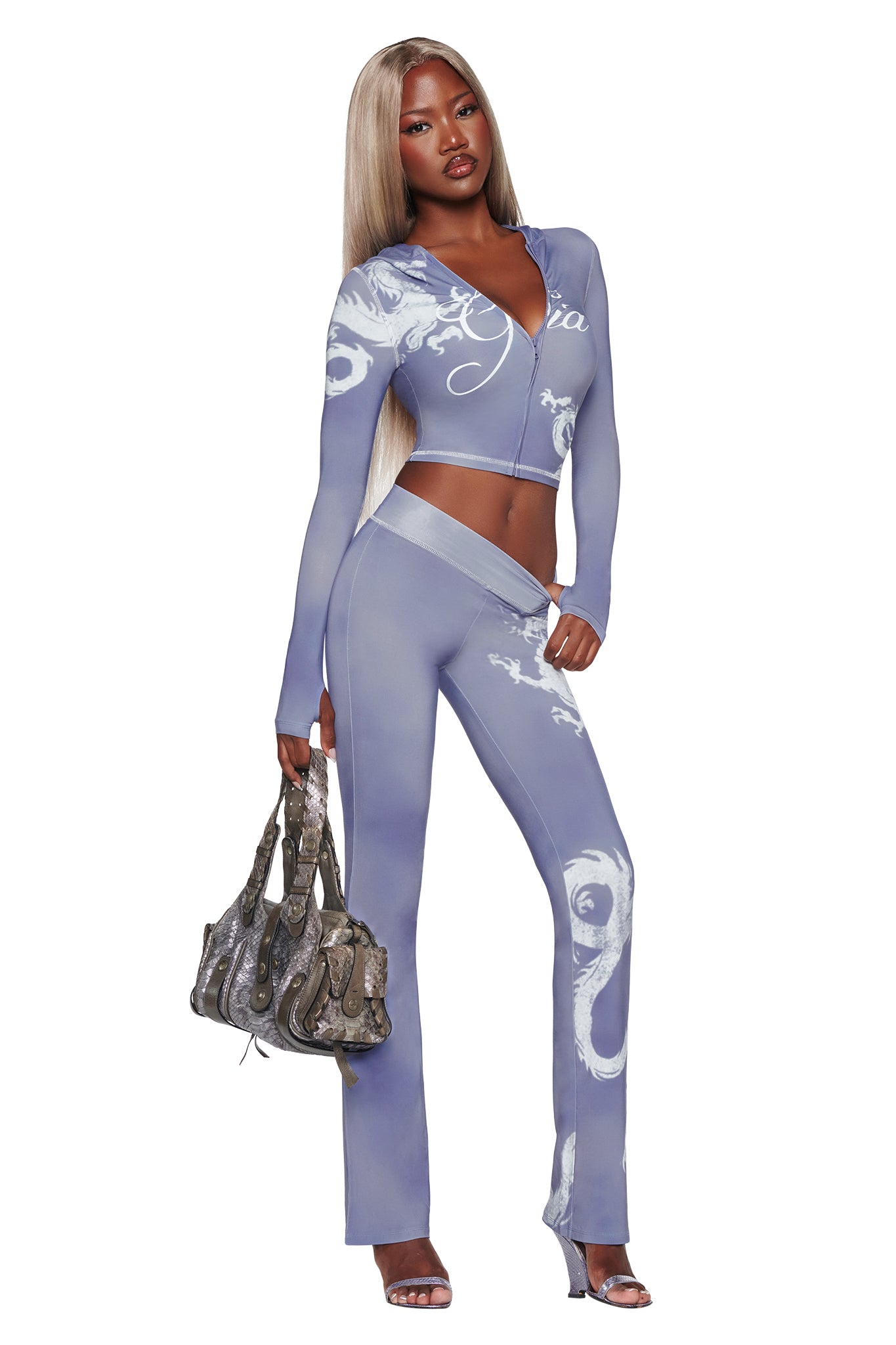 A woman with long blonde hair stands confidently against a white background, wearing a fitted lavender two-piece outfit with white patterns. Draped casually over her shoulders is the I.AM.GIA BLARE HOODIE in Steel Blue Dragon. She completes the look with a textured handbag and heeled sandals.