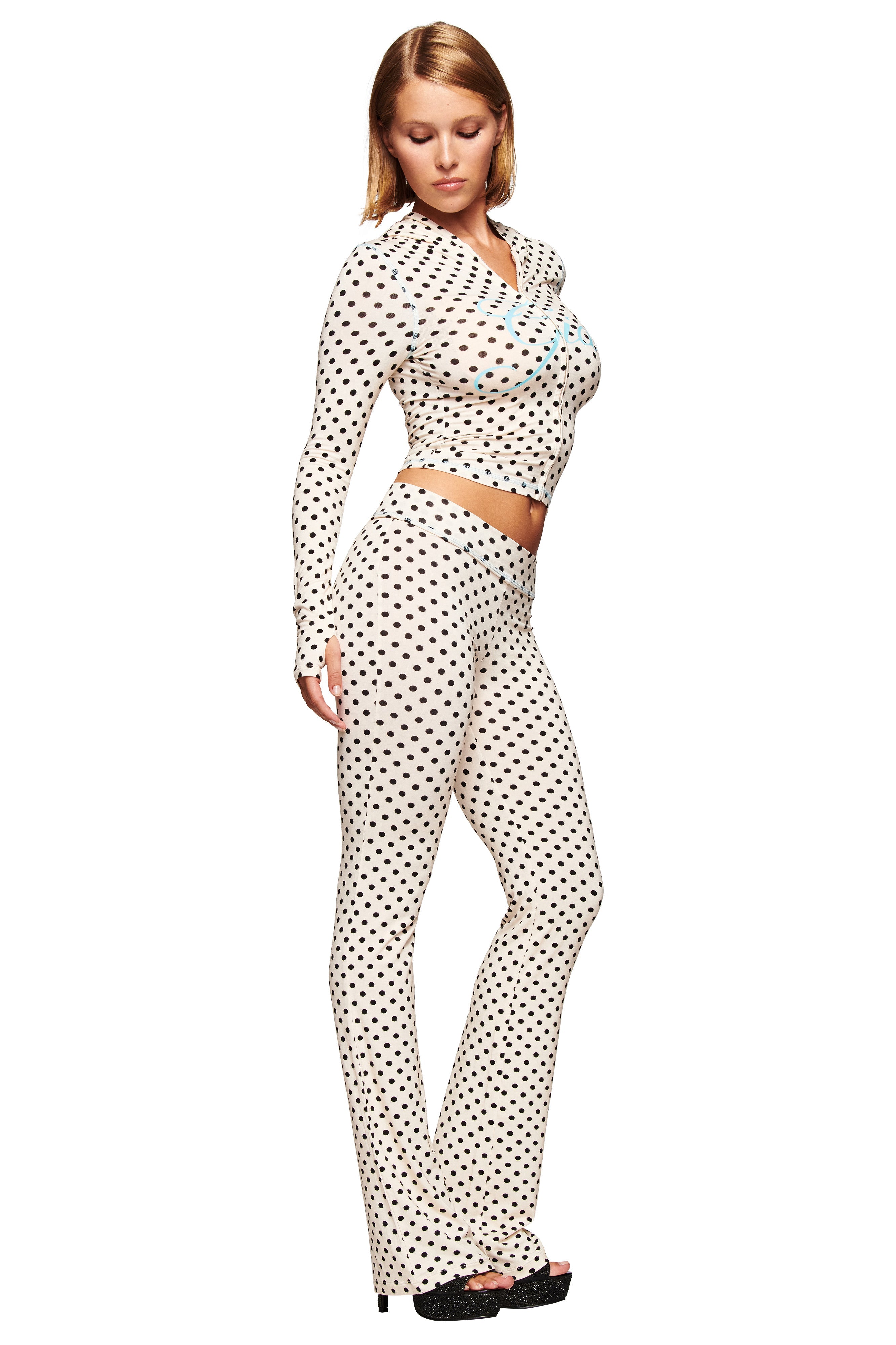 A woman poses in the I.AM.GIA BLARE HOODIE in Cream Polka Dot, styled with matching high-waisted pants and black platform shoes against a minimalist white background. She looks downward, showcasing the striking polka dot print.