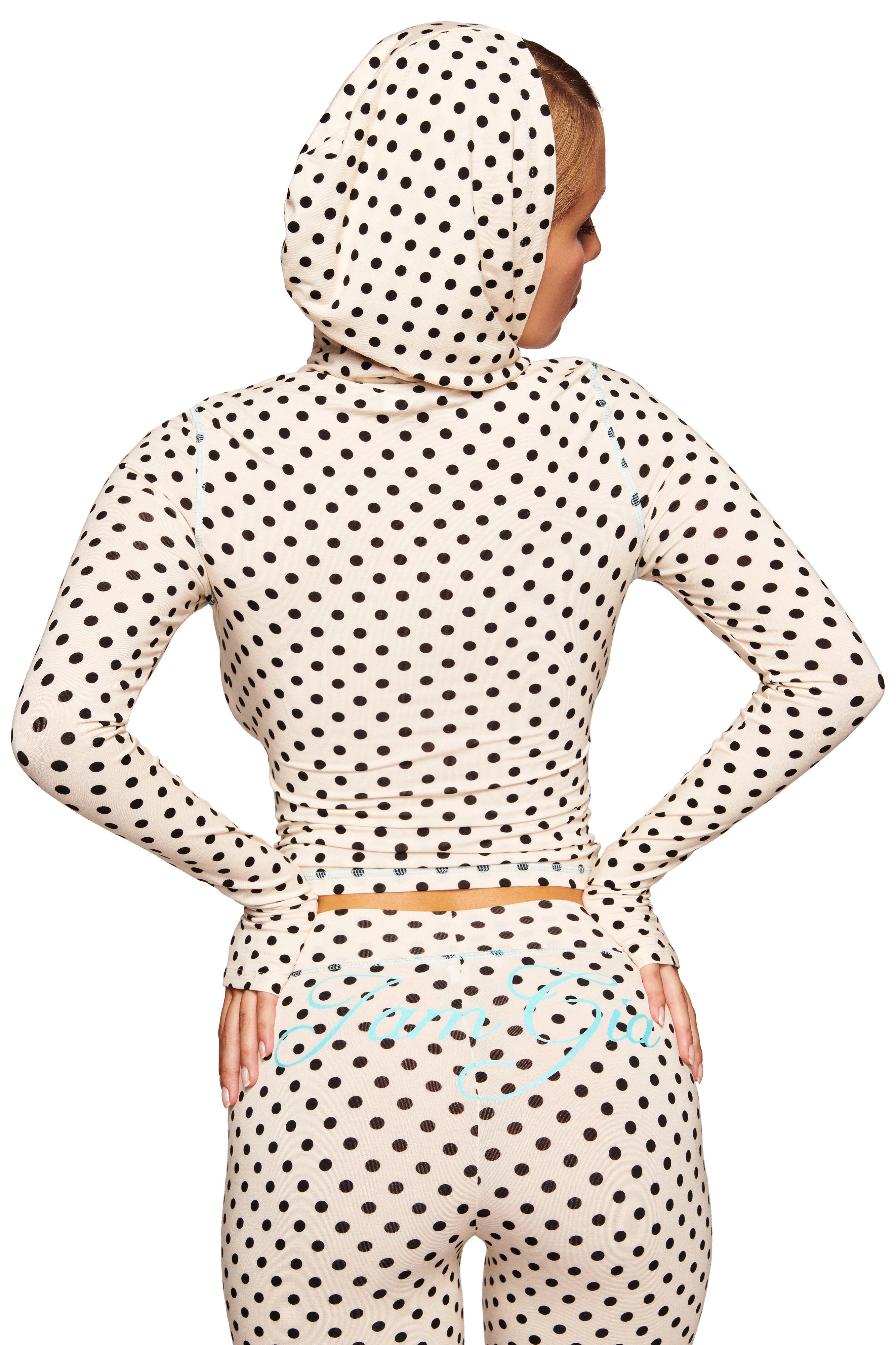 A woman faces away, hands on hips, wearing I.AM.GIA’s BLARE TRACKPANT in cream polka dot—a flared leg, long-sleeve hooded outfit with a playful print and the hood up, partly covering her hair.