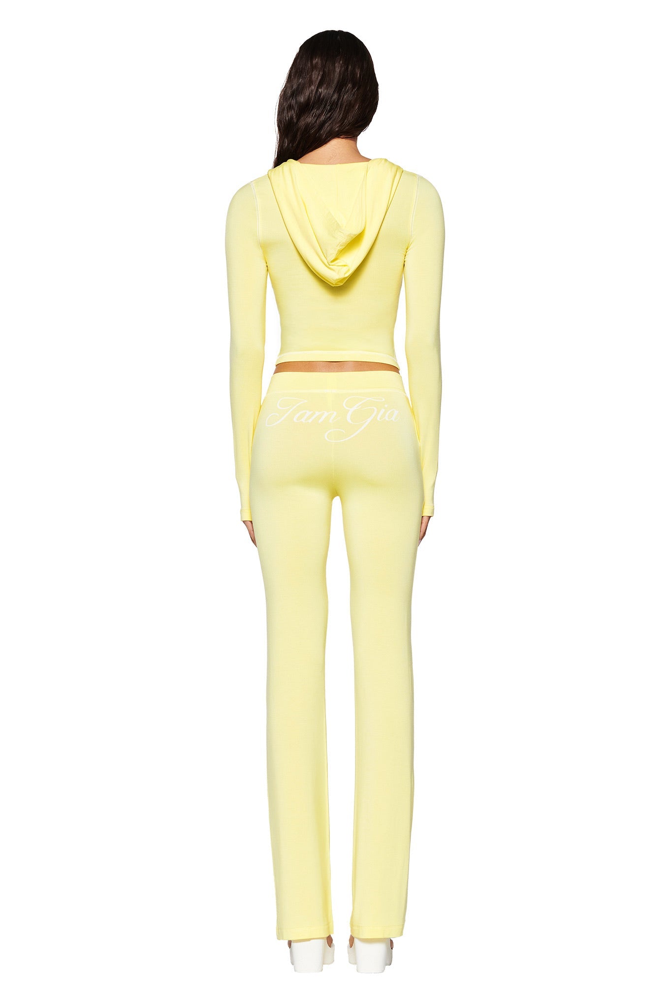 A person with long hair stands facing away, wearing a form-fitting pale yellow Blare Trackpant and hooded top by I.AM.GIA. The outfit, crafted from bamboo and jersey materials, showcases the bold "I.AM.GIA" logo on the back against a pristine white background.