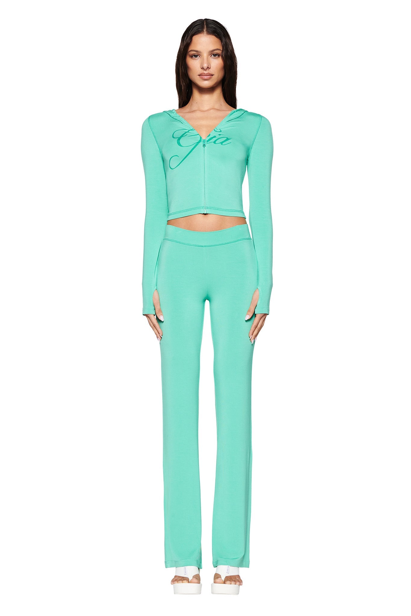 A person poses neutrally against a white background, wearing I.AM.GIA's turquoise teal long-sleeve crop top with "GCDS" printed on it and matching high-waisted Blare Trackpant in Green: Turquoise: Teal, paired with open-toed beige heels.