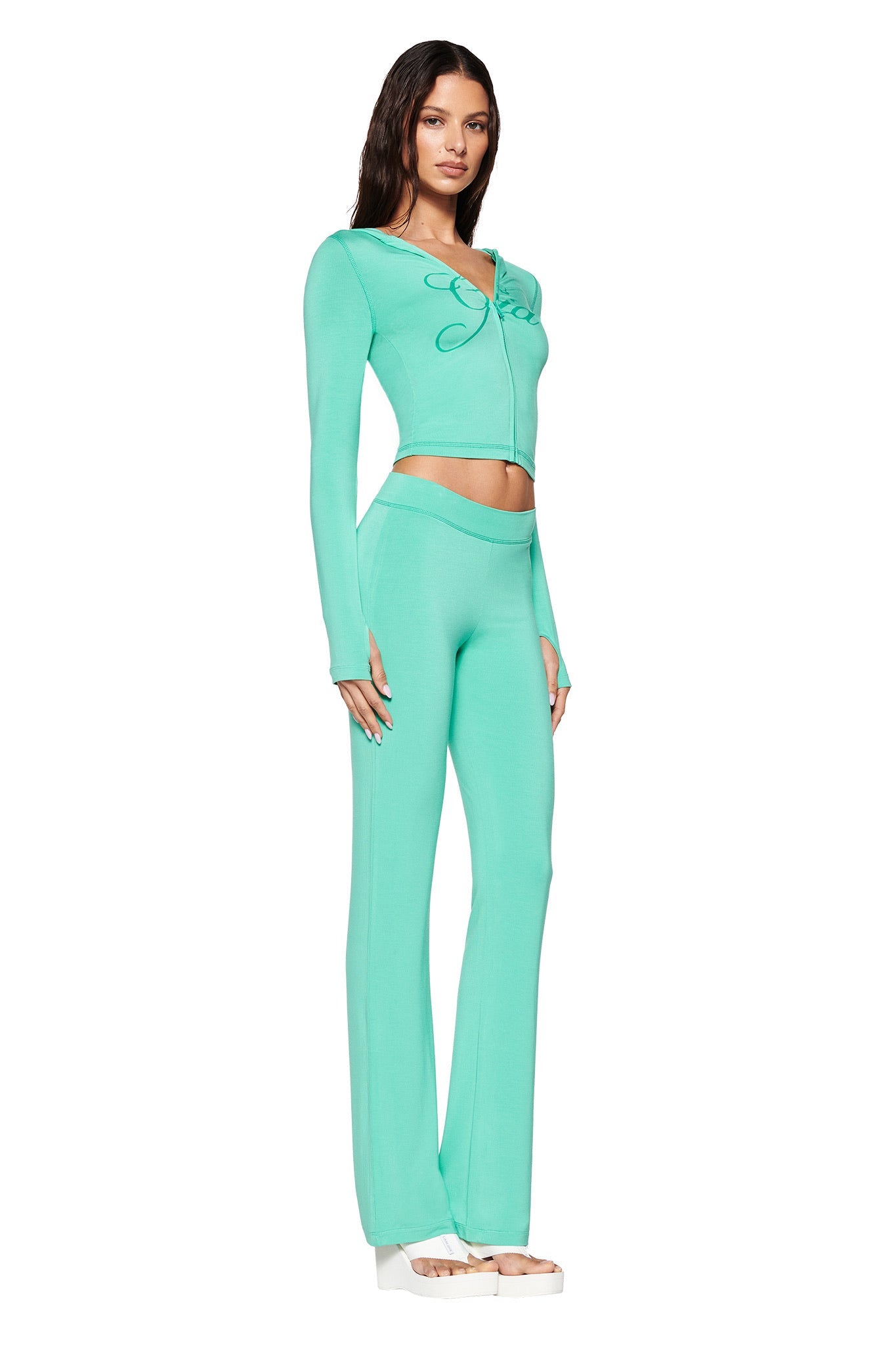 A woman poses in a three-quarter view on a white background wearing I.AM.GIA's BLARE TRACKPANT in turquoise teal with a tied crop top and platform sandals. The outfit, made from soft, stretchy bamboo fabric, pairs comfort with style.