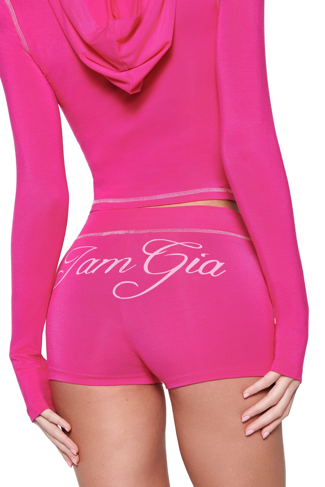 A person confidently wears a bright pink I.AM.GIA hoodie paired with the BLARE SHORT - PINK, showcasing a matching bodycon fit and "Tam Gia" stylishly displayed across the back. Their right hand rests on their left hip.