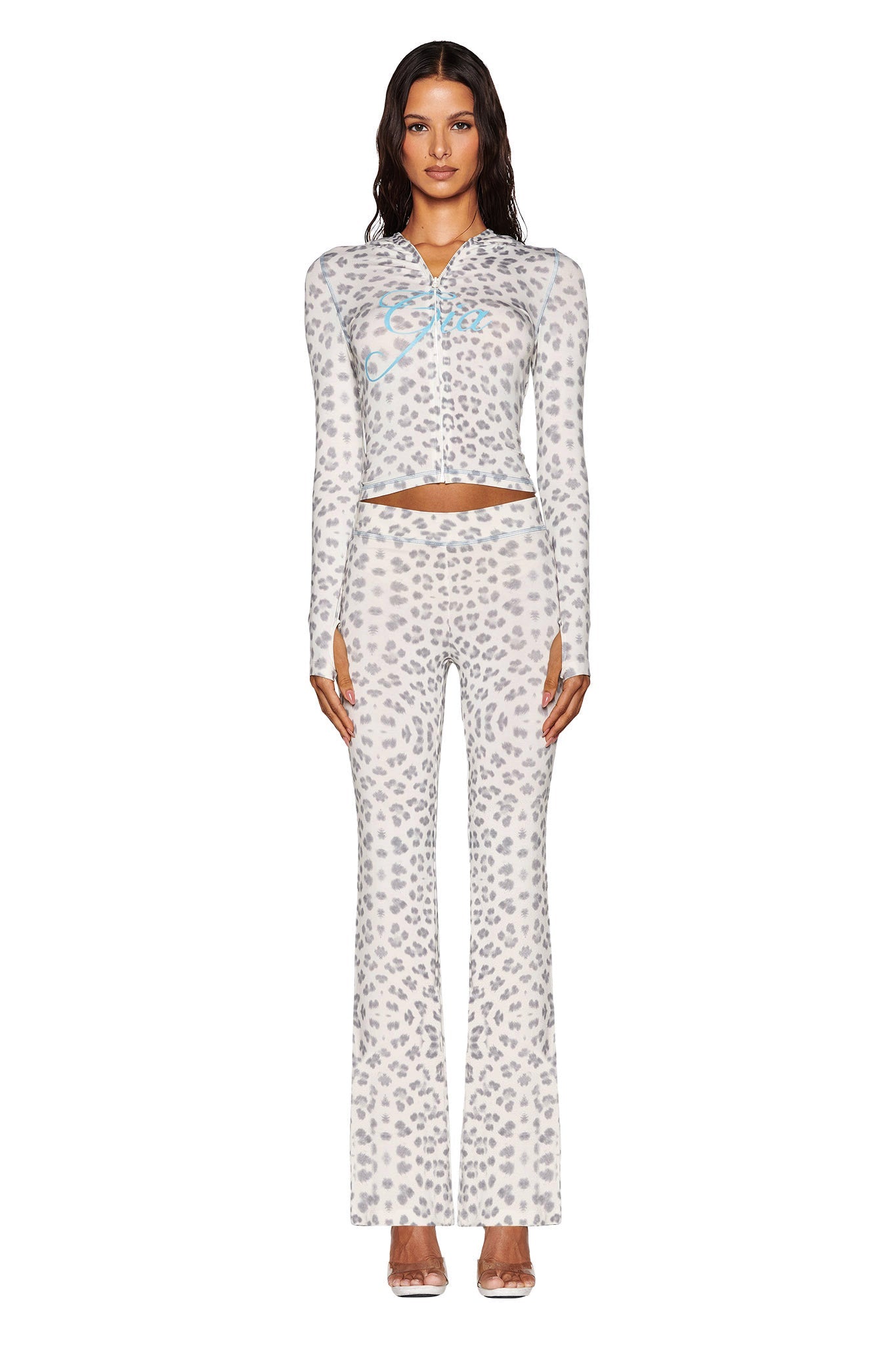 A person stylishly wears the I.AM.GIA BLARE TRACKPANT from the Snow Leopard: Winter Wild Card collection, along with a matching long-sleeve top. The elegant light gray snow leopard print on a white background complements their long, wavy hair and transparent high heels, exuding a chic, modern aesthetic.