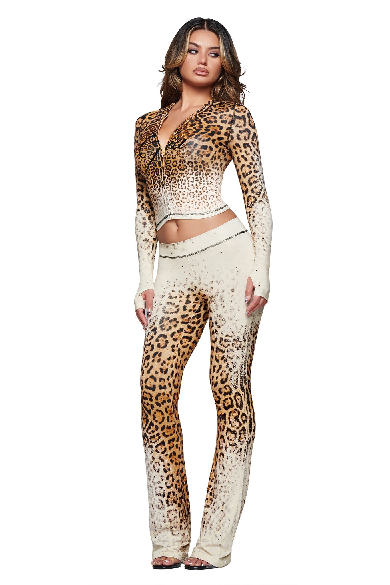 A woman stands confidently against a white background, wearing the I.AM.GIA BLARE TRACKPANT in Cream Faded Panthera—a fitted, long-sleeve leopard-print crop top paired with matching cheetah print track pants. Her hair falls in loose waves and she completes the look with open-toe heels.