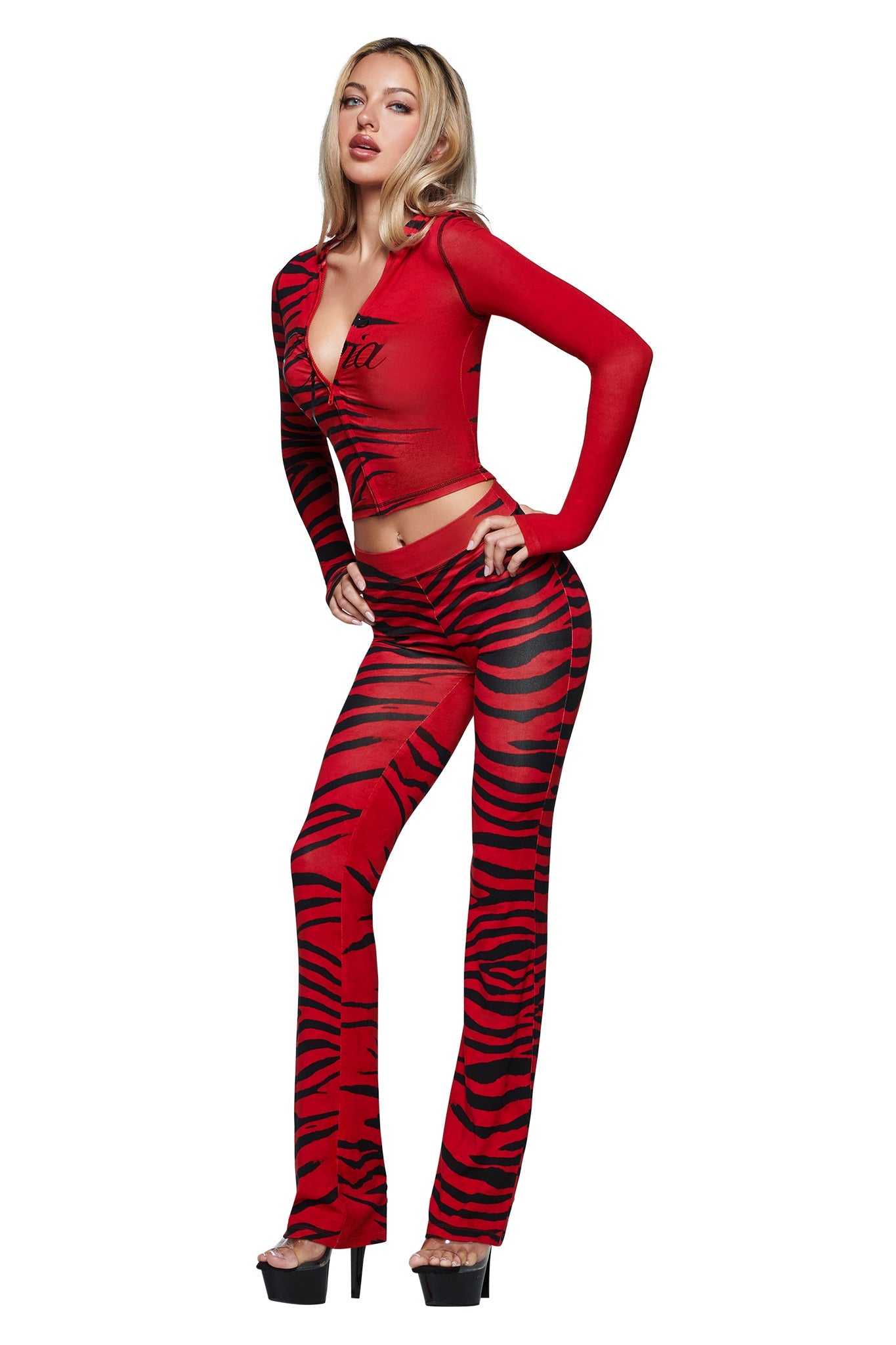 A woman wears the I.AM.GIA BLARE TRACKPANT - RED WILD SIDE high-waisted pants with a matching red, sheer, tiger-striped crop top and black platform heels. She stands confidently with one hand on her hip, looking at the camera.