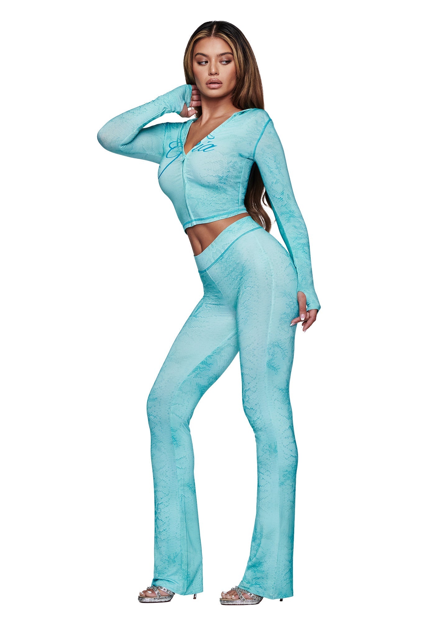A woman models the I.AM.GIA BLARE HOODIE in teal serpentine with matching high-waisted flared pants. She pairs the look with silver open-toed heels and wears her long, straight hair against a white background.