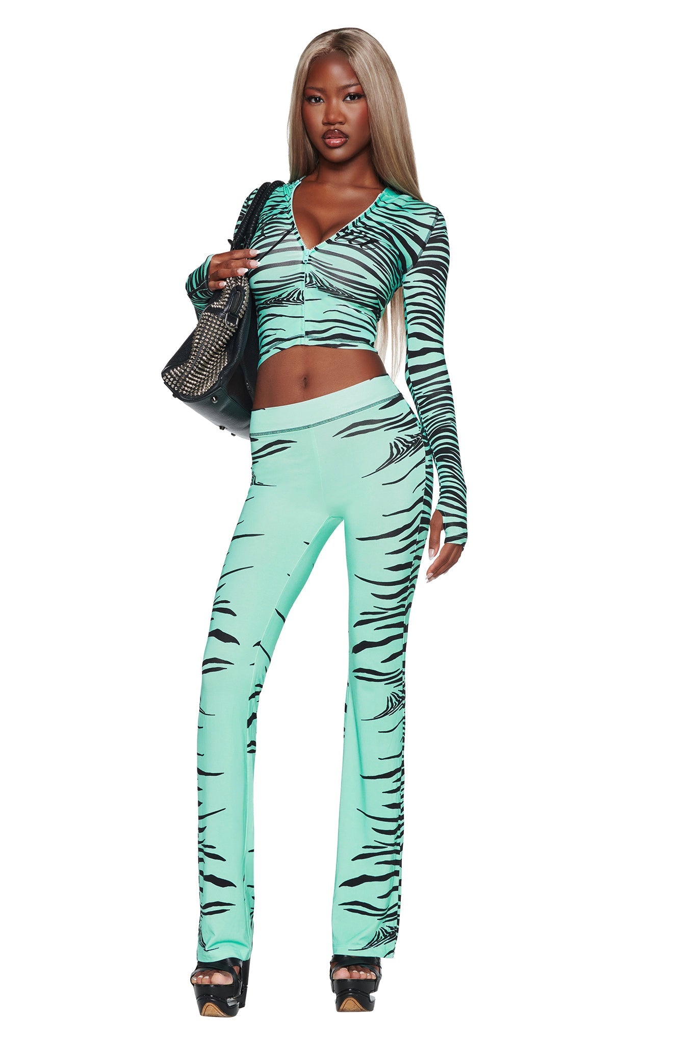 A woman with long blonde hair wears the BLARE TRACKPANT - AQUA FINE LINE ZEBRA by I.AM.GIA, paired with a matching turquoise and black tiger-striped crop top, black heels, and a black bag. She stands confidently against a plain white background.