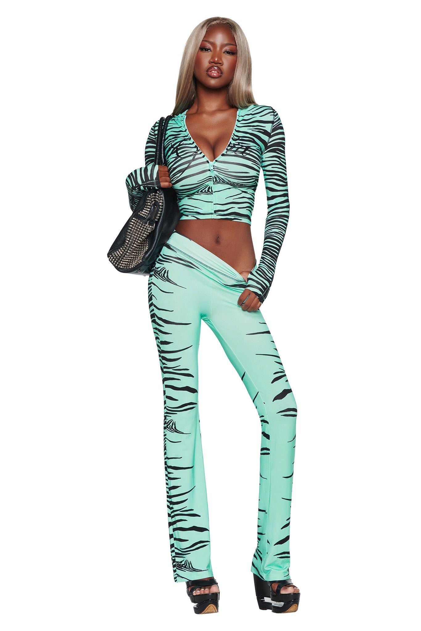 A woman with long, straight, blonde hair models the I.AM.GIA BLARE TRACKPANT in Aqua Fine Line Zebra, featuring a bold aqua and black zebra print with a stylish flared leg. She pairs it with a coordinating crop top, black platform sandals, and a black studded handbag as she poses confidently against a white background.