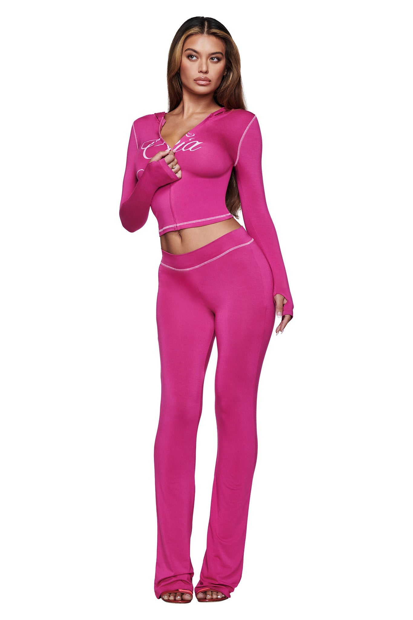 A woman with long brown hair and heeled sandals wears the I.AM.GIA BLARE TRACKPANT in Raspberry with white trim, paired with a bright pink fitted long-sleeve top featuring white script text.