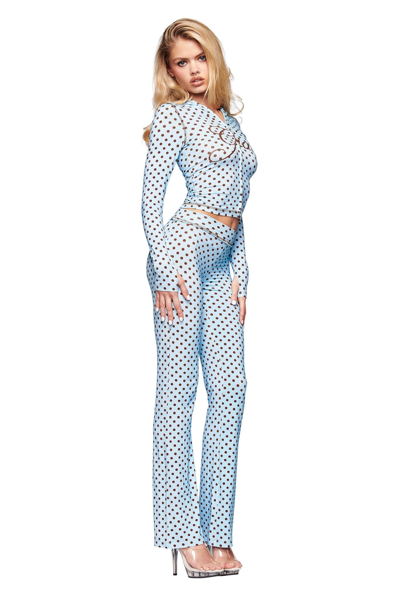 A woman poses in the I.AM.GIA BLARE TRACKPANT - BLUE POLKA DOT, a sheer, long-sleeve light blue outfit with black polka dots and low-rise waist. She wears clear heels, her long blonde hair loose, against a white background.