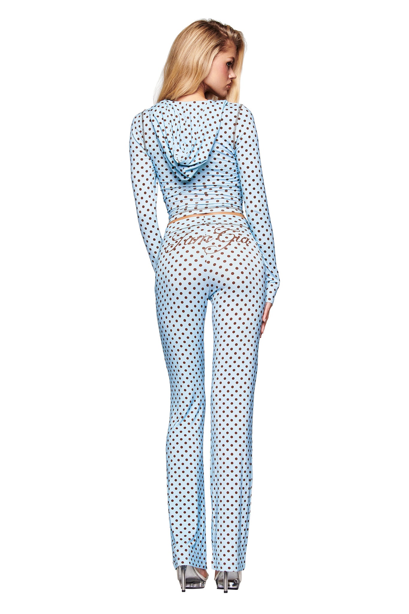 A woman faces away wearing I.AM.GIA's BLARE TRACKPANT - BLUE POLKA DOT, a light blue hooded mesh outfit with black polka dots, low-rise waist, silver heels, and cursive text on the back.