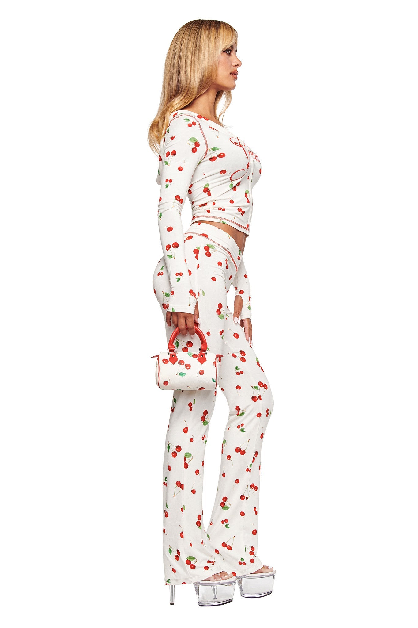 A woman stands in profile wearing I.AM.GIA's BLARE HOODIE - CHERRY ON TOP, paired with flared pants, clear platform heels, and holding a small matching purse.