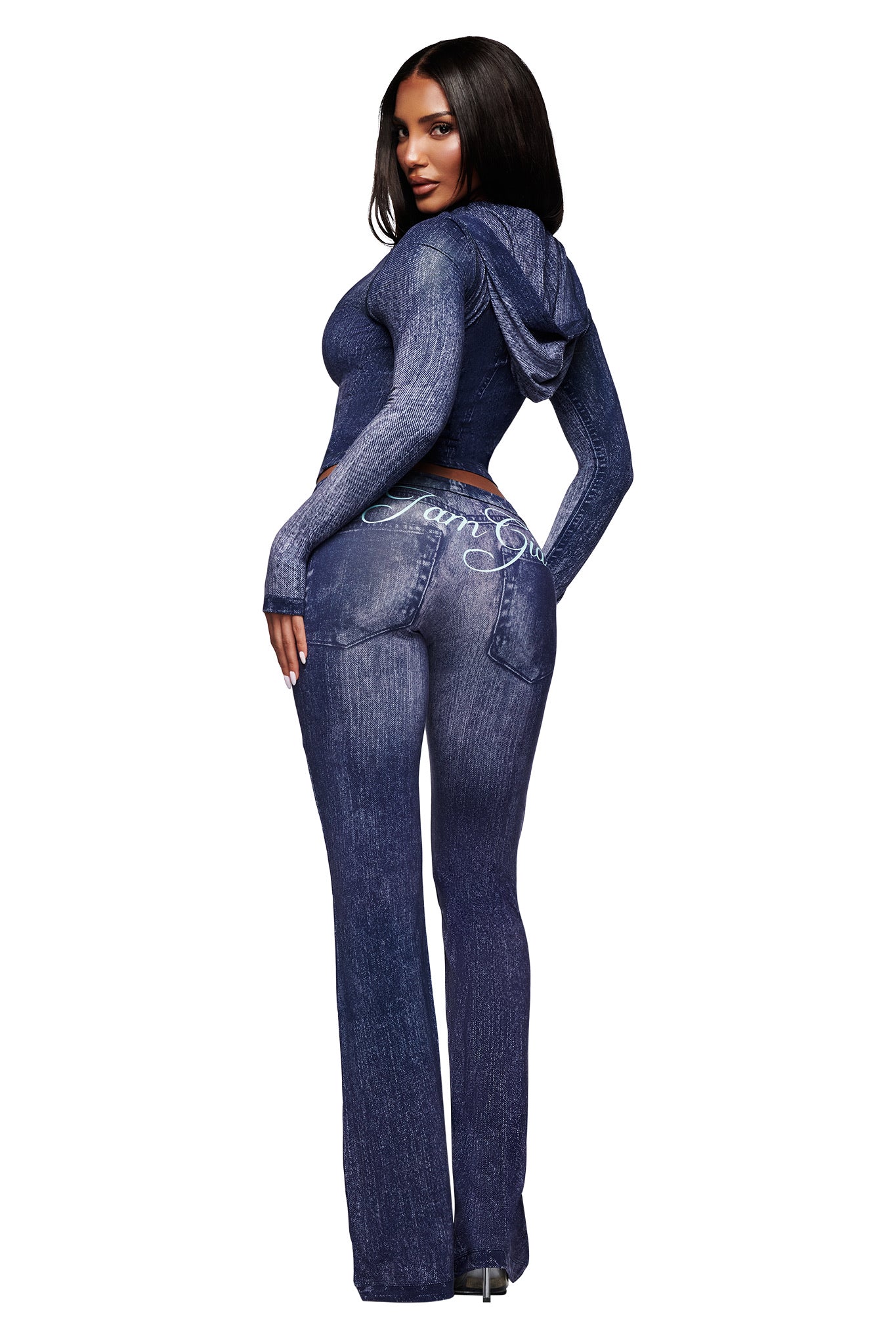 A woman with long dark hair poses facing backward, wearing I.AM.GIA's BLARE TRACKPANT - DARK DENIM, a fitted faux-denim trackpant featuring "Sami S" in white script on the back pocket. She stands against a white background.