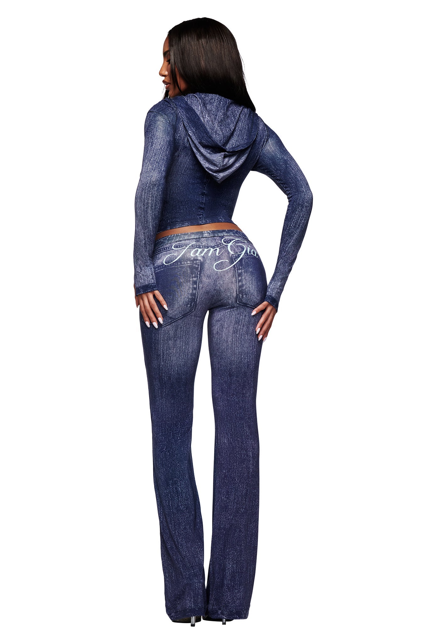 A woman with long dark hair wears the I.AM.GIA BLARE HOODIE - DARK DENIM and matching fitted pants, standing hands on hips. "I am Gia" is scripted in white on the back of her pants.