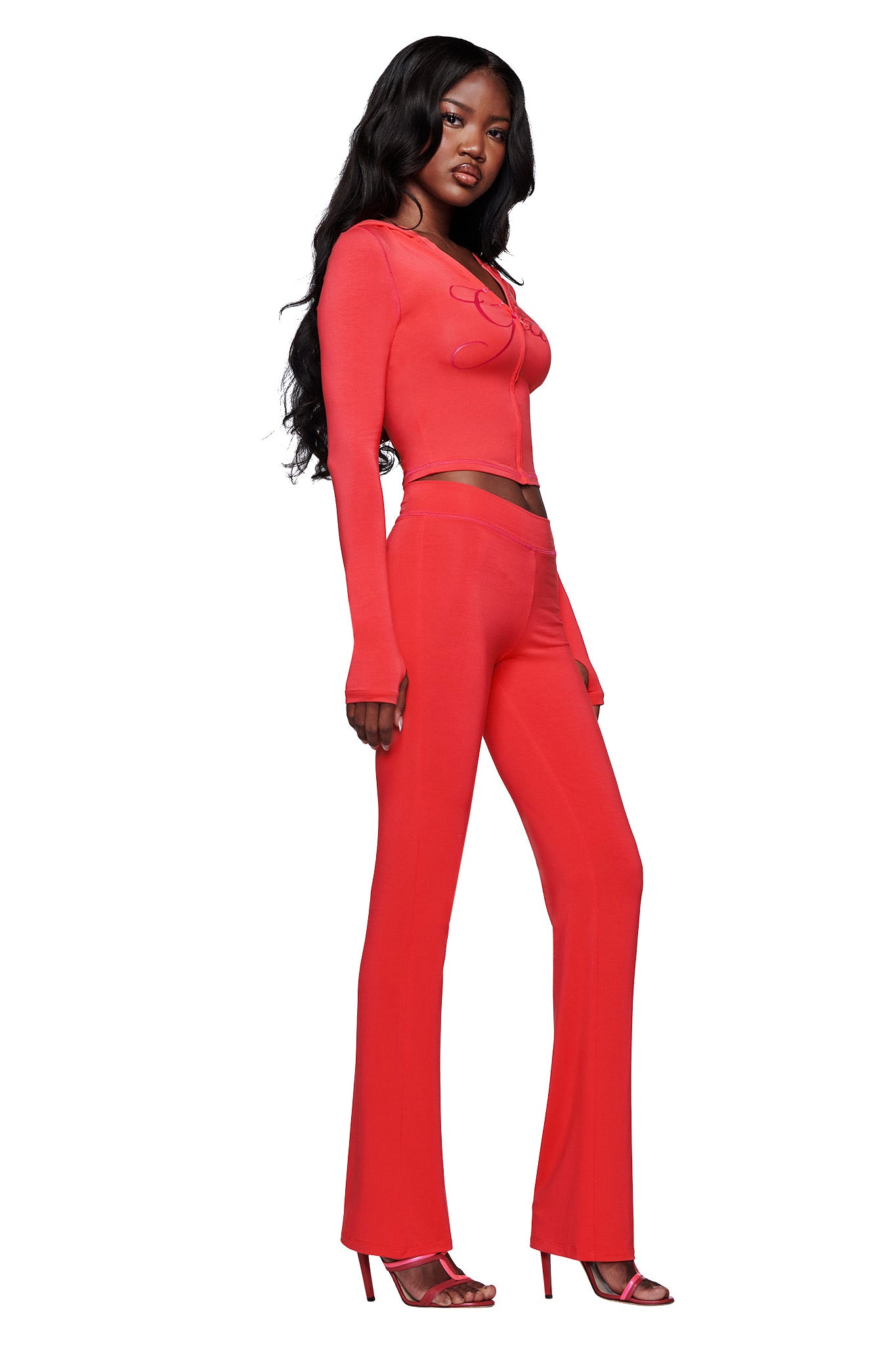 A woman with long, wavy black hair poses against a white background wearing I.AM.GIA’s BLARE TRACKPANT in Papaya—a bright red, fitted, mid-rise flare pant—styled with a matching long sleeve top and red heels.