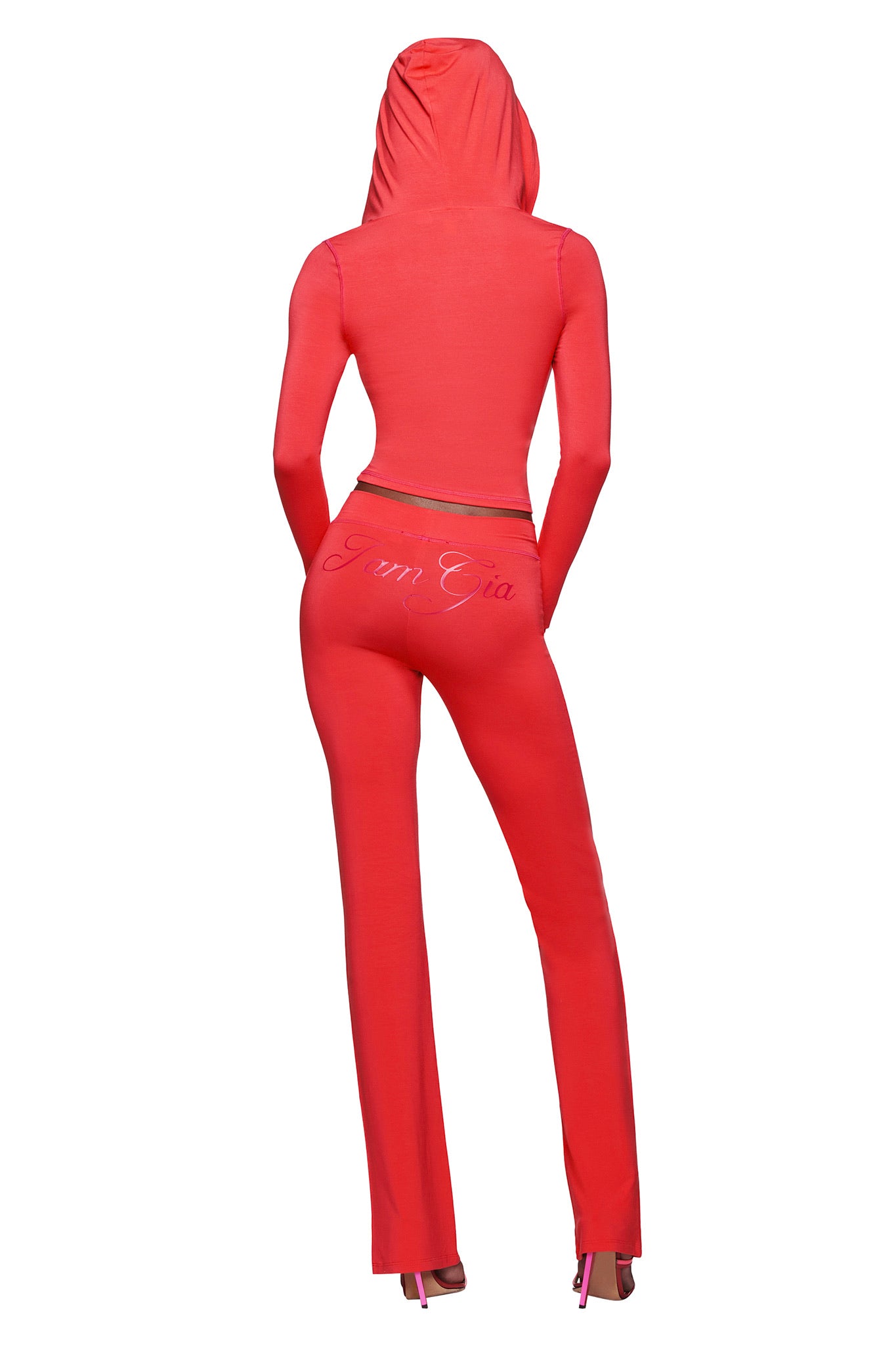 A person faces away in a fitted red hooded top and BLARE TRACKPANT - PAPAYA by I.AM.GIA, featuring "I Am Gia" on the back. The pants’ mid-rise waist and flared leg pair with pink heels, all set against a white background.
