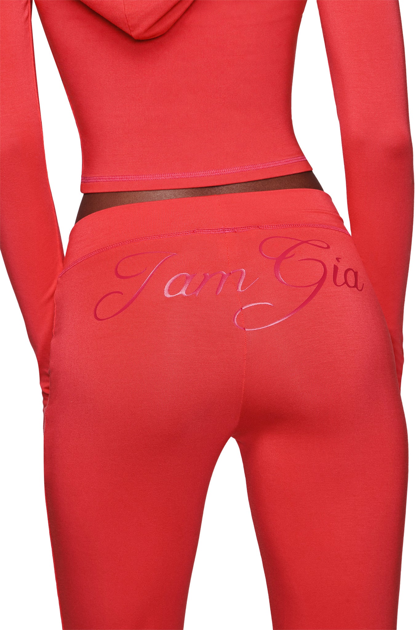 A person wears the I.AM.GIA BLARE TRACKPANT in Papaya, featuring a mid-rise waist with "IamGia" in shiny pink script across the back waistband and flared legs for a trendy look.