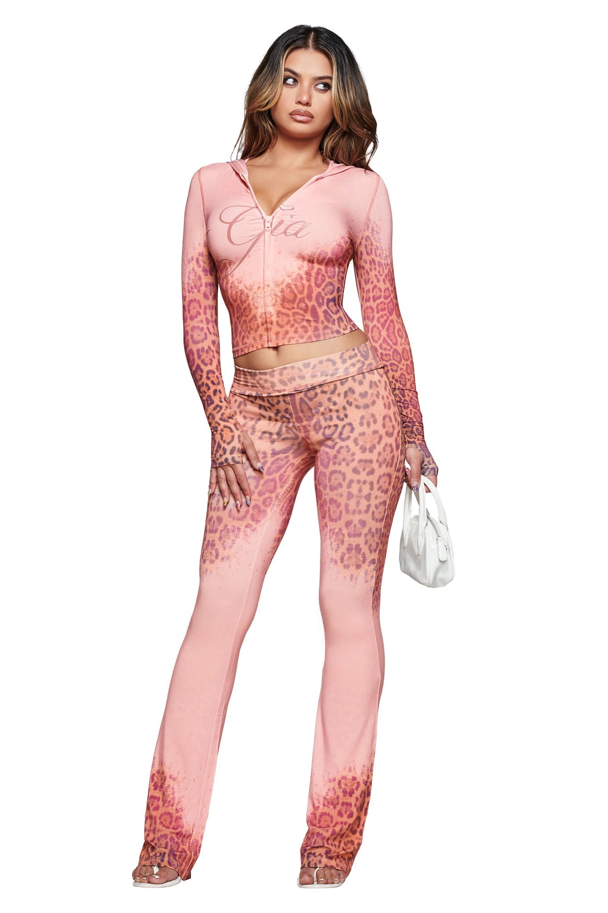 A woman stands and poses in the I.AM.GIA BLARE TRACKPANT - PINK FADED PANTHERA trackpants as part of a pink panthera print two-piece outfit, holding a small white purse with her long loose hair, against a white background.