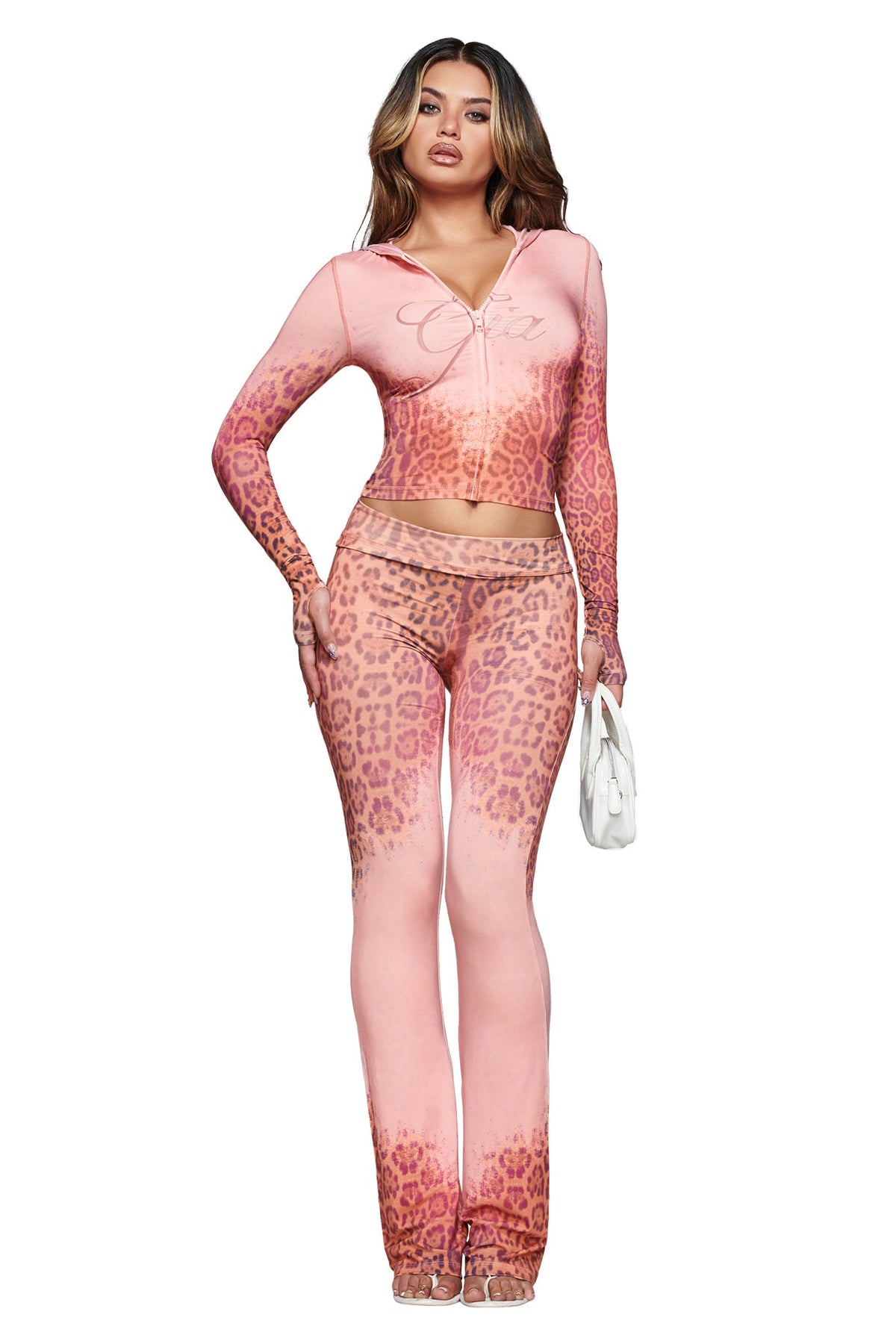 A woman poses against a white background wearing the I.AM.GIA BLARE TRACKPANT - PINK FADED PANTHERA in a matching panthera print set with long sleeves and flared pants, accessorized with a small white handbag.