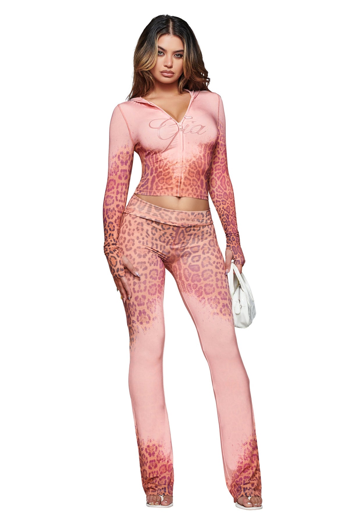 A woman stands against a white background, wearing the I.AM.GIA BLARE TRACKPANT in Pink Faded Panthera with a matching long-sleeve top, and holds a small white handbag.