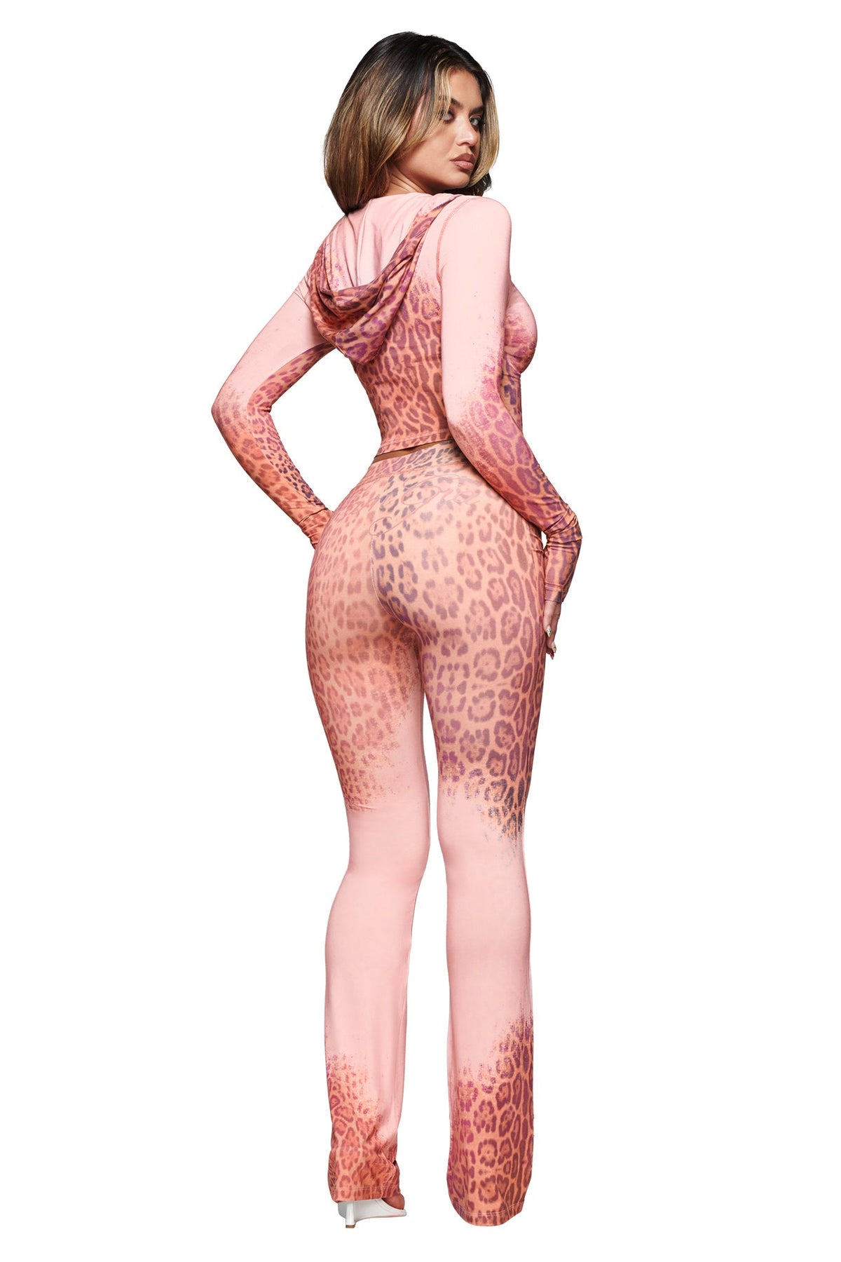 A woman stands facing away, wearing the BLARE HOODIE - PINK FADED PANTHERA by I.AM.GIA—a fitted, pink leopard-print jumpsuit with long sleeves, flared legs, and a hood—posing against a white background.