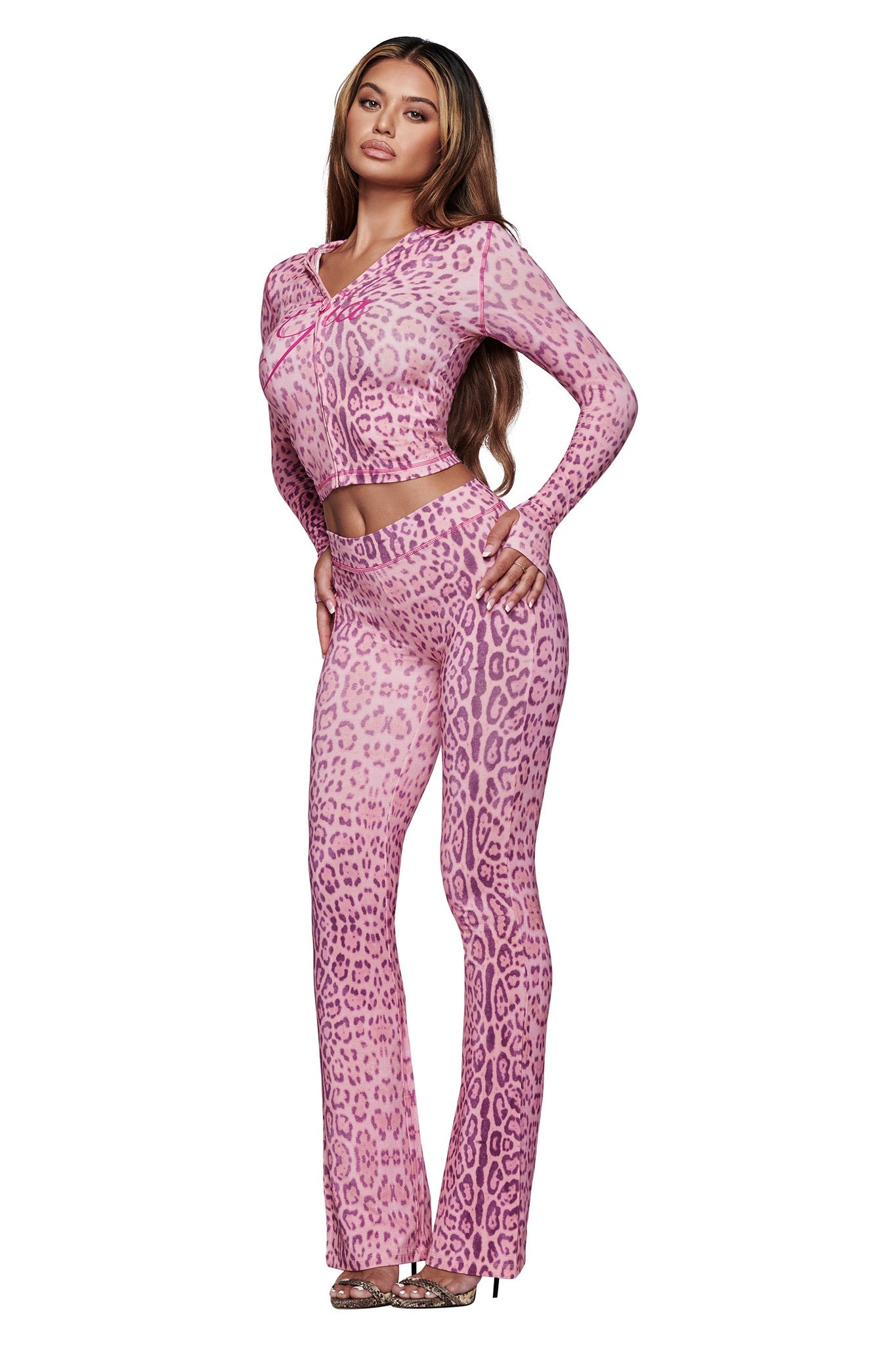 A woman poses in I.AM.GIA’s BLARE TRACKPANT - PINK PANTHERA, a high-waisted, flared-leg trackpant paired with a matching long-sleeve crop top. She has long brown hair and open-toe heels, standing against a plain white background.