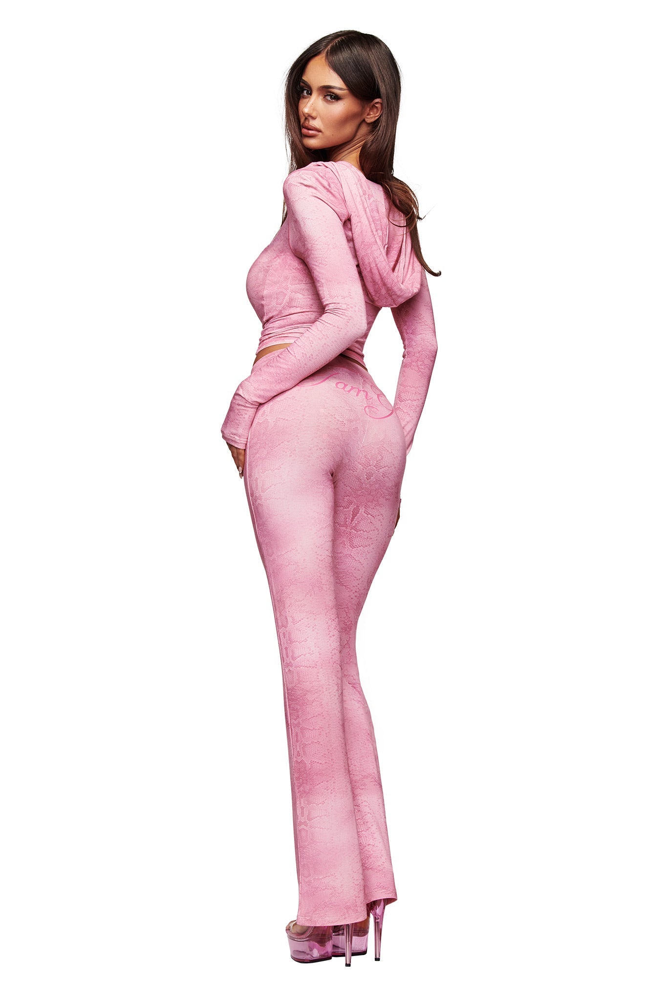 A woman stands on a plain white background, glancing over her shoulder in a fitted pink hooded top and I.AM.GIA's BLARE TRACKPANT - LIGHT PINK SERPENTINE with a flared leg, paired with platform heels.