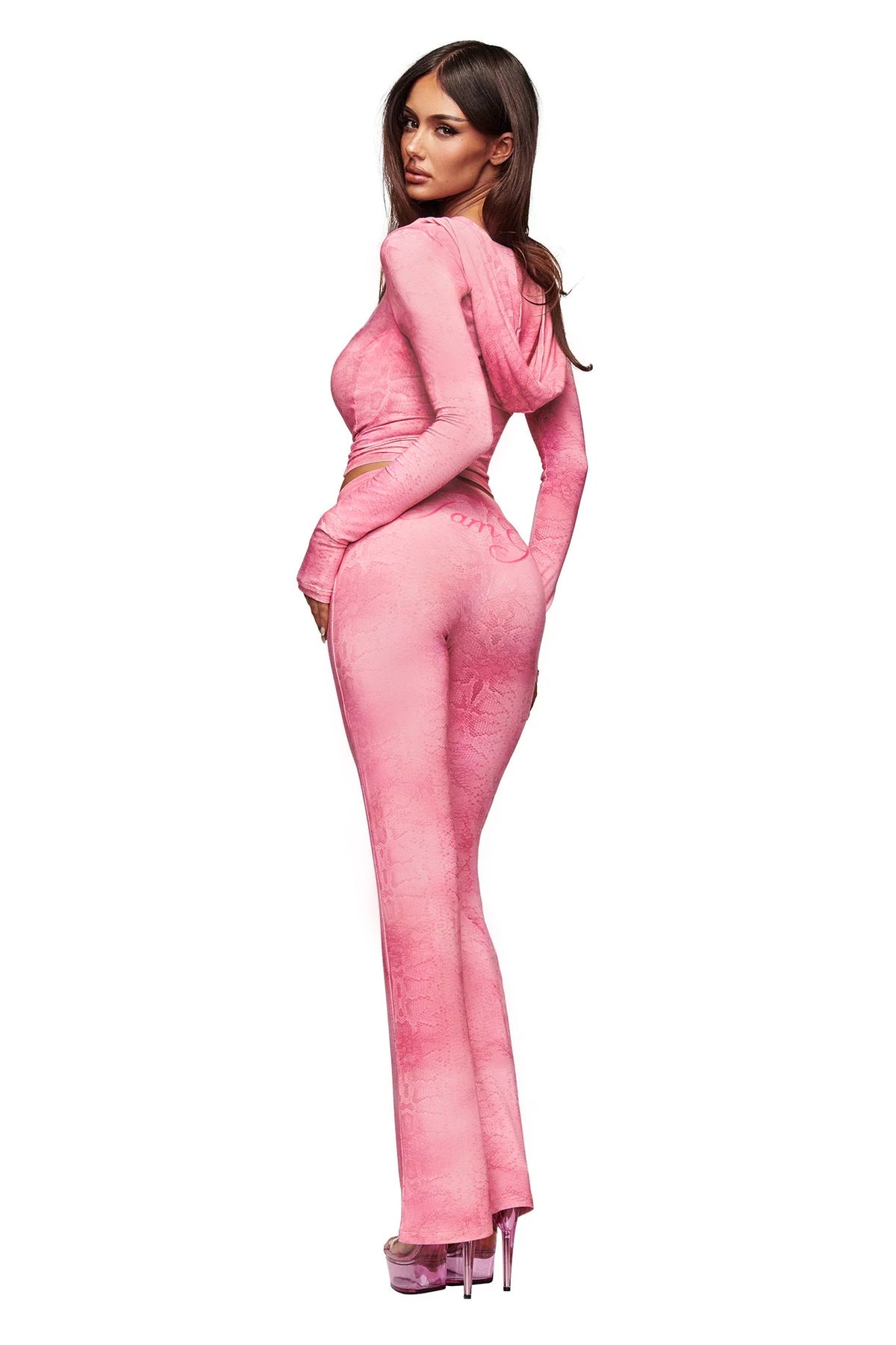 A woman wearing the I.AM.GIA BLARE TRACKPANT in Light Pink Serpentine, styled with a fitted, long-sleeve hooded top and platform heels, stands and looks over her shoulder against a white background.