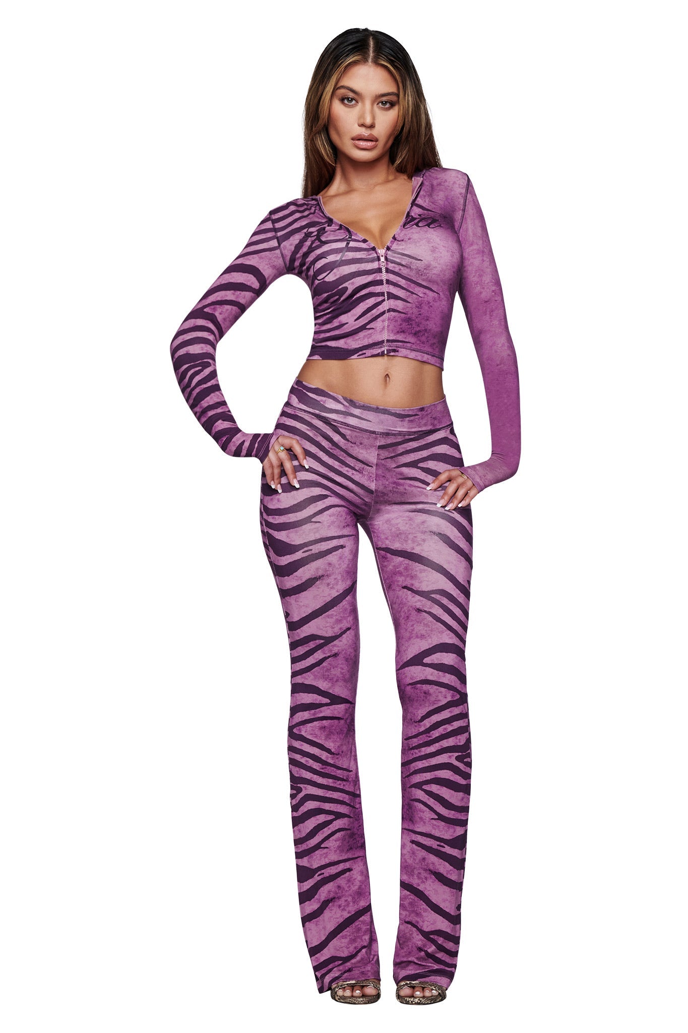 A woman faces forward wearing the I.AM.GIA BLARE HOODIE in Plum Wild Side zebra print with long sleeves, paired with matching flared pants and clear heels. She has long brown hair and stands against a white background.