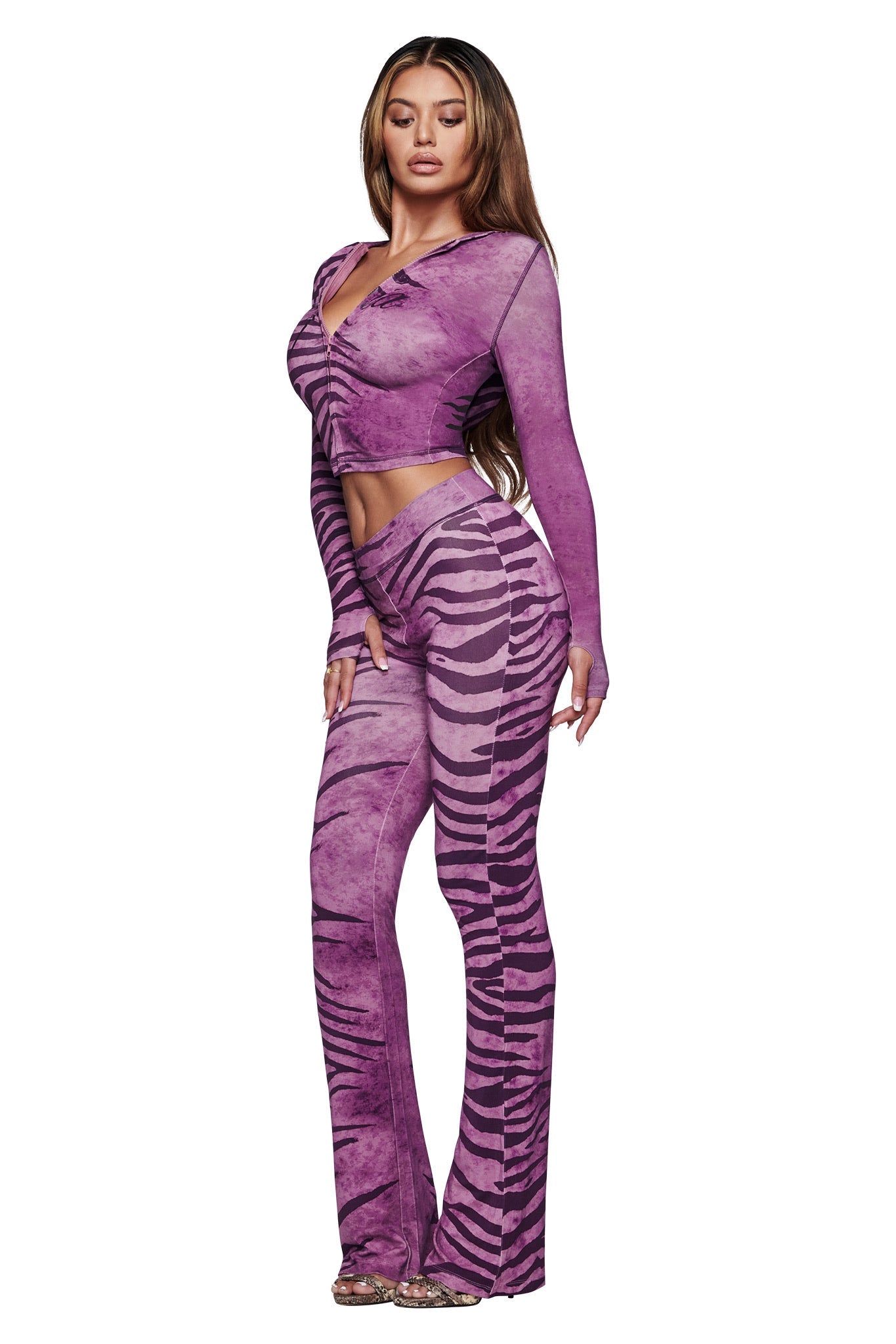 A woman models the I.AM.GIA BLARE HOODIE in Plum Wild Side, showcasing its bold purple zebra print. She pairs it with clear high heels and stands against a white background.